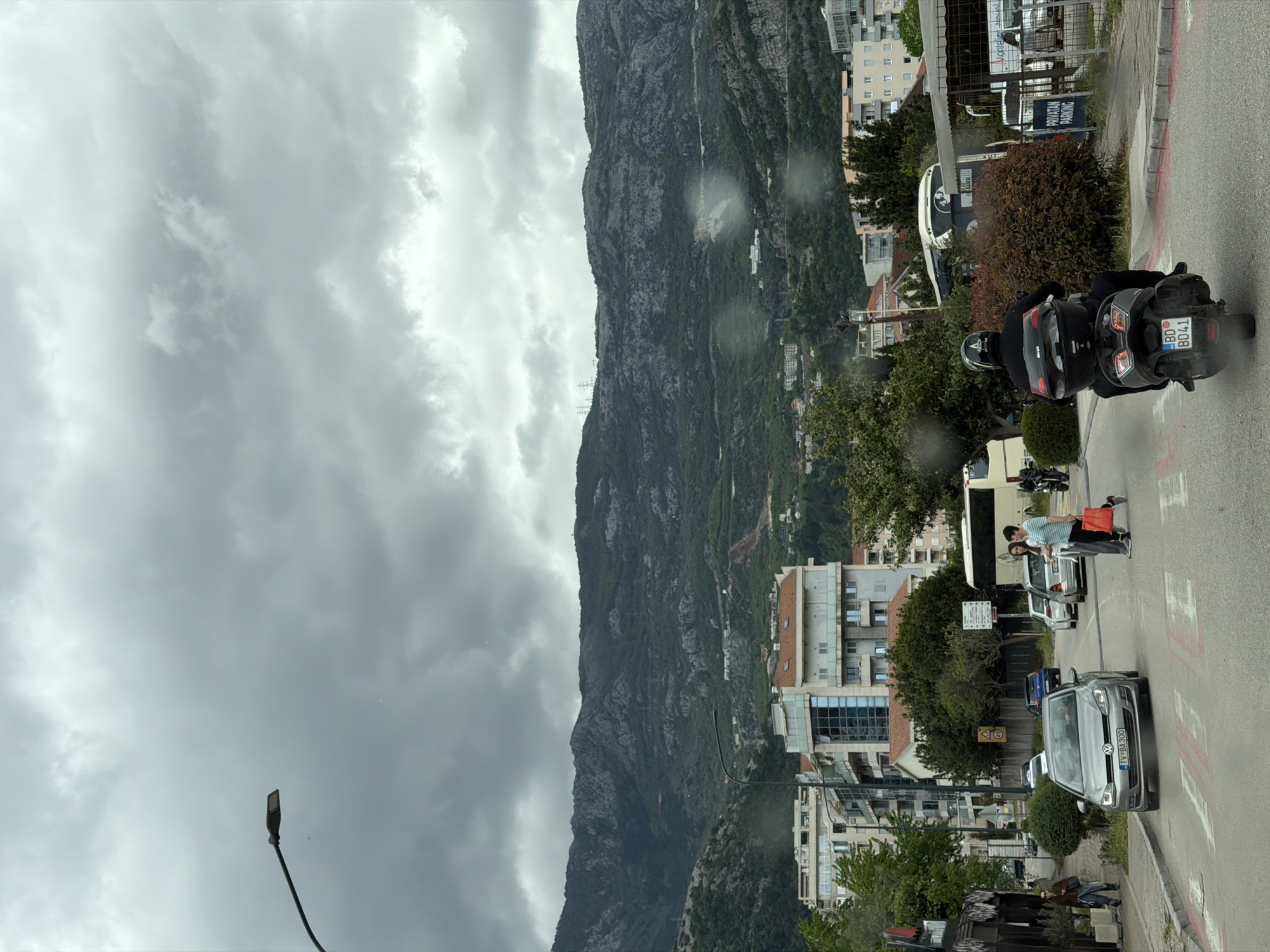 The image presents a street scene in Budva, Montenegro, under a heavily overcast sky. A massive, rugged mountain, partially covered in dense vegetation, dominates the background, rising steeply behind the urban development. The photograph appears to have been taken from inside a vehicle, evidenced by water droplets or smudges visible on what looks like a window or windshield.
In the immediate foreground, a person is seated on a black motorcycle, viewed from behind. The rider wears a helmet and dark clothing, and the motorcycle is equipped with GIVI brand luggage. Its license plate clearly displays "BD 8041," with "BD" indicating Budva, Montenegro. Further down the road, a silver Volkswagen Golf, bearing the Montenegrin license plate "TV BA300" (TV for Tivat), approaches. Two pedestrians are visible crossing the street further ahead, one carrying a vibrant red bag. A white bus is partially obscured by a large bush on the right side of the road.
The street is lined with multi-story buildings, characteristic of a Mediterranean or Balkan town, interspersed with trees and bushes. A tall street lamp stands on the left side of the frame. Visible text includes the license plates "BD 8041" and "TV BA300," the "GIVI" logo on the motorcycle luggage, and a sign behind the bus that reads "PRIVATAN PARKING" (Private Parking). The overall impression is a cloudy daytime scene of everyday life and traffic within a mountainous coastal setting.