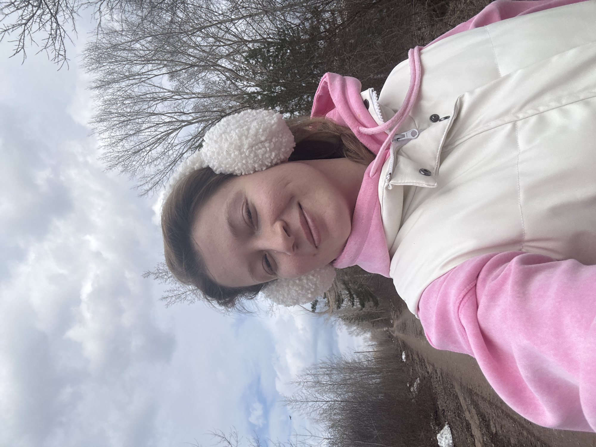 A young woman is captured in a selfie-style portrait taken outdoors, with the image rotated 90 degrees counter-clockwise. She is positioned towards the center-left of the frame, looking directly at the camera with a subtle, content smile. Her light brown hair is visible, and she is wearing warm attire including a bright pink hoodie under a white puffer vest. Distinctive white, fluffy earmuffs cover her ears.
The background primarily features an overcast, cloudy sky. Beneath the sky and behind the woman, numerous bare trees with intricate branches dominate the landscape, indicative of a late autumn, winter, or early spring setting. To the right and lower portion of the image, a dirt path or trail is visible, with small patches of white that appear to be snow or ice, reinforcing the cool weather conditions. The scene suggests a serene, chilly day outdoors in what is contextually Chaikovsky, Russia. No text is visible in the image.