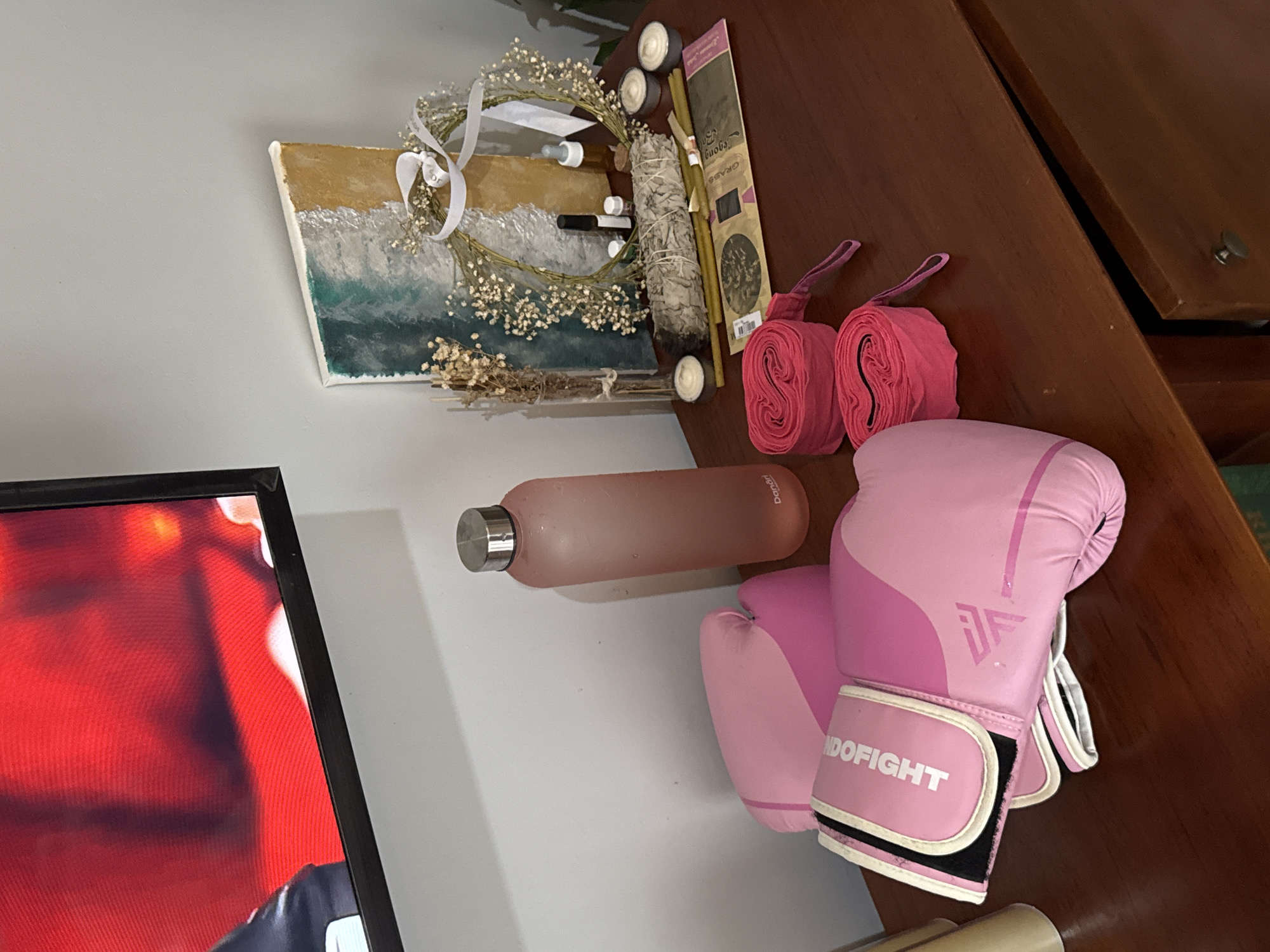 A close-up shot captures a collection of boxing and spiritual items arranged on a dark wood dresser in North Kuta, Indonesia. In the foreground, two pink boxing gloves with white trim are stacked, one resting on top of the other. Next to them, two rolled-up pink hand wraps are neatly positioned. To the right, a tall, frosted pink water bottle with a silver cap stands upright.
Behind the gloves and water bottle, a decorative display includes a small canvas painting featuring abstract ocean and beach imagery. A delicate wreath adorned with small white flowers and a white ribbon is placed in front of the painting. Several thin, green candles are arranged in a row, along with a bundle of white sage, and a package of incense sticks. Small glass bottles, likely containing oils or perfumes, are also part of this arrangement. The scene is illuminated by soft, ambient lighting, and a portion of a television screen displaying a bright red image is visible in the upper left corner. The overall impression is one of a personal space dedicated to both physical training and spiritual well-being.