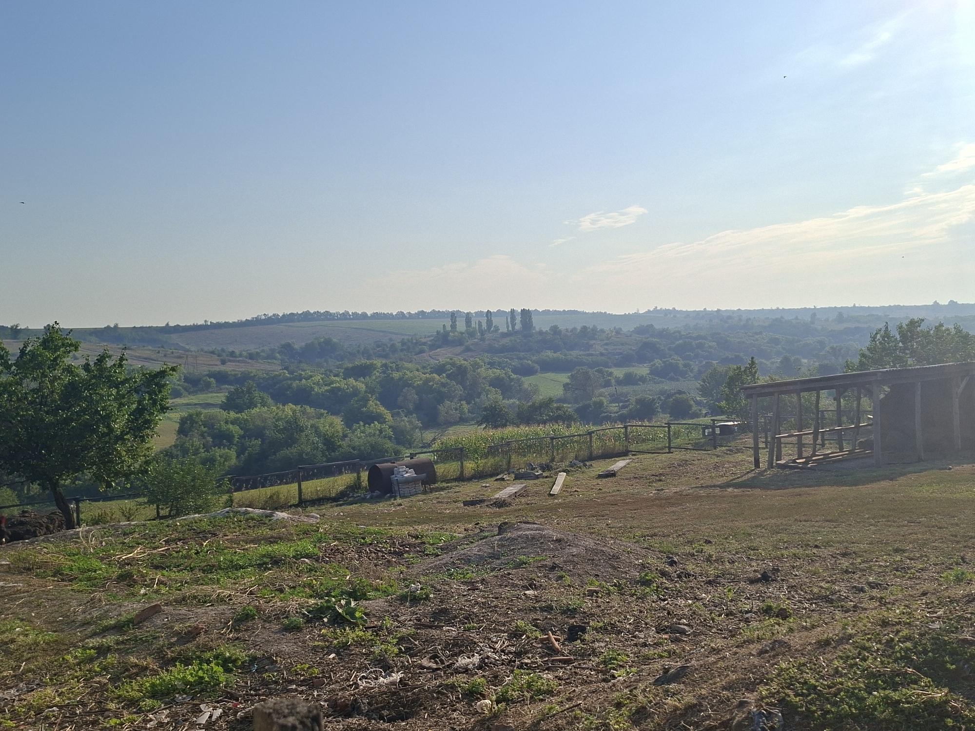This is a wide-angle, eye-level photograph taken in Oleksandropil, Ukraine, featuring a rural landscape under a clear blue sky with scattered clouds. The foreground is rough, uncultivated ground with scattered debris, dry grass, and some patches of green vegetation. A large pile of dirt or sand is visible on the right side of the foreground.
Moving back, a wooden fence runs horizontally across the midground, separating the immediate foreground from a slightly lower, more cultivated area. Beyond the fence, there are further patches of green fields, some with visible rows of crops, and dense clusters of trees. In the far distance, rolling hills covered in trees and vegetation extend to the horizon.
On the right side of the image, a simple wooden open-sided structure with a sloped roof is present. It appears to be a shelter or possibly a simple storage area, with wooden beams and supports visible. To the left of this structure, the ground slopes down, and a dark, barrel-like object sits near the fence, with some stacked concrete blocks beside it. A large, leafy tree stands prominently on the left side of the midground. The overall impression is of a pastoral, possibly agricultural setting on a bright, sunny day. No people are visible in the frame.