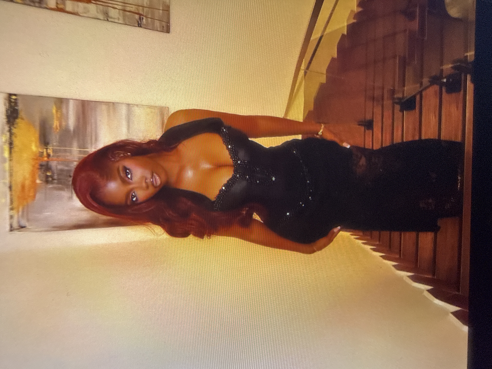 A Black woman with long, wavy red hair stands on a wooden staircase. She is wearing a sleeveless, floor-length black dress with a fitted bodice adorned with rhinestones. The dress has a plunging neckline and a halter top. Her left arm is bent, resting on her hip, and she wears a gold bracelet on her wrist. The staircase has dark wooden steps and a railing. Behind her, on the wall, are two abstract paintings with warm, golden tones. The overall lighting suggests an indoor setting. There is no explicit indication of this location being in Abuja, Nigeria.