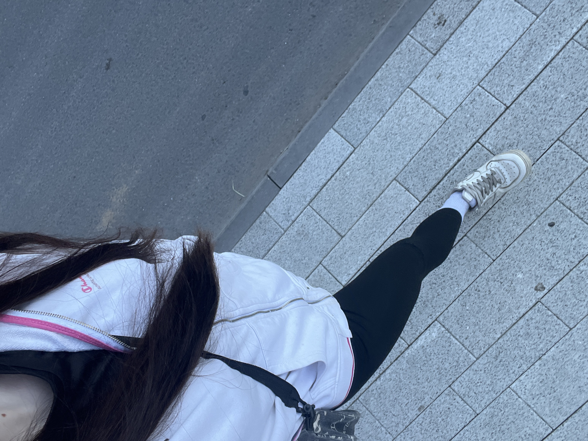 This overhead image captures a person walking or standing on a street in Saint Petersburg, Russia, with a focus on their lower body and the ground. The individual wears black leggings, white socks, and white athletic shoes with grey laces and light beige accents.
Their left side is partially visible, showing long, dark hair falling over a white jacket or hoodie. This outerwear features a prominent pink lining along its silver zipper. A black top is worn beneath the jacket, and a black strap, likely from a bag, crosses their torso. A small logo on the white jacket is partially visible, appearing to read "Champions" or "Champion" with "Authentic Athletics" underneath, set within a circular design.
The ground is segmented into two distinct areas. On the left, a dark grey, textured surface resembles asphalt, indicative of a road or pathway. To the right, a sidewalk is paved with light grey, rectangular interlocking stones arranged in a linear pattern. A dark grey curb acts as a clear demarcation between these two surfaces. The lighting suggests it is daytime, with diffused light indicating an overcast sky or soft ambient conditions, as no harsh shadows are present. The scene portrays a common urban outdoor environment.