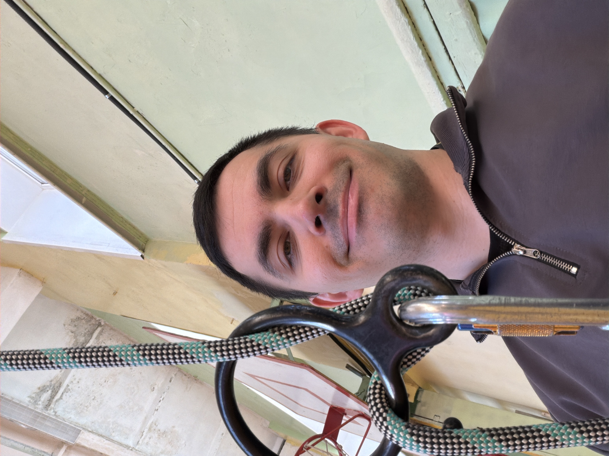 A man is suspended from a rope. The rope is black and green. The carabiner and climbing equipment are attached to the rope. The man is wearing a gray jacket with a zipper. The location is unknown.