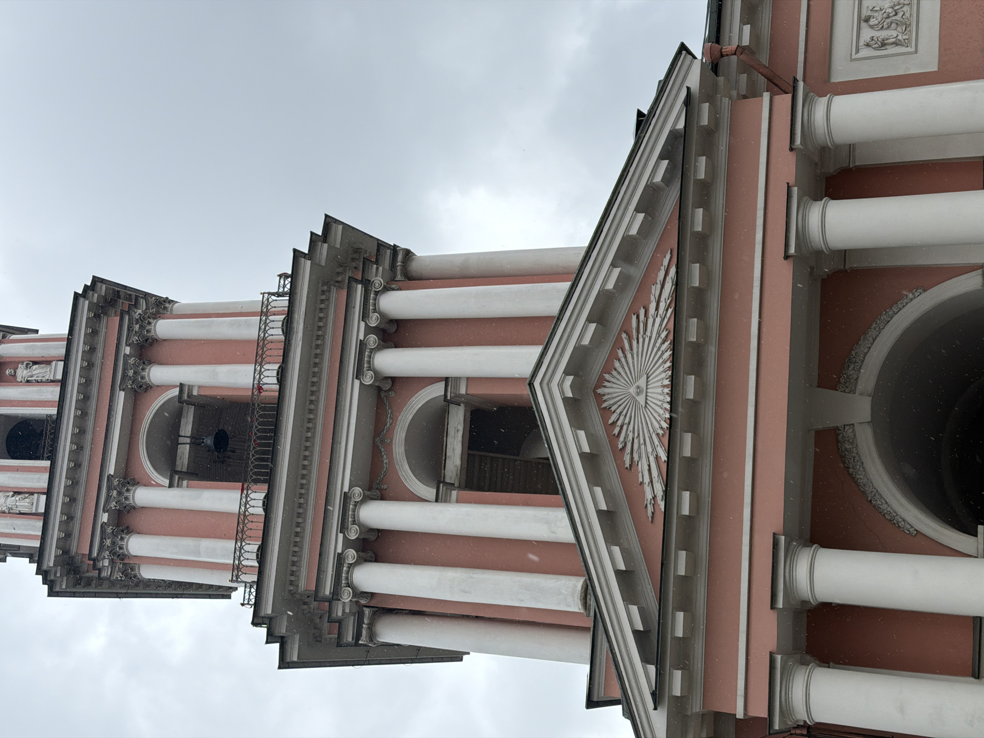The image shows a low-angle view of the bell tower of the Smolny Cathedral in Saint Petersburg, Russia. The architecture is predominantly Baroque, featuring pastel pink and white colors, with columns, arched openings, decorative moldings, and ornamentation. The tower is multi-tiered, with each level decreasing in size as it rises. A decorative sunburst design is visible on the facade of one of the tiers. The sky is overcast, and snow is falling. A bell is visible inside one of the arched openings.