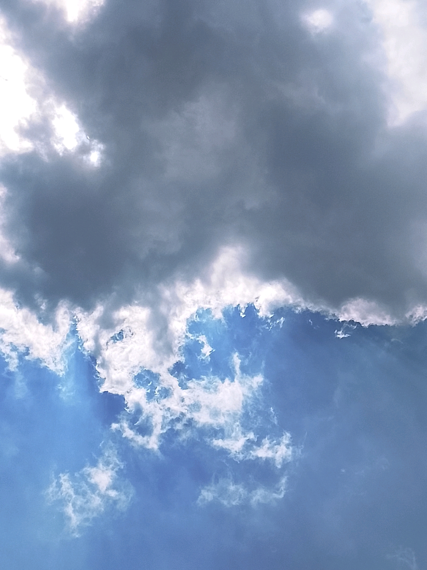 The image displays a sky with dramatic cloud formations. The upper half is dominated by dark, heavy storm clouds, with patches of bright white light breaking through. The lower half of the sky is a vivid blue, dotted with scattered, wispy white clouds. The transition between the dark and light clouds is sharp, creating a contrast that suggests changing weather. There are no visible buildings, people, or identifiable landmarks that would place the scene in Slawi, Indonesia. The image focuses solely on the sky.
