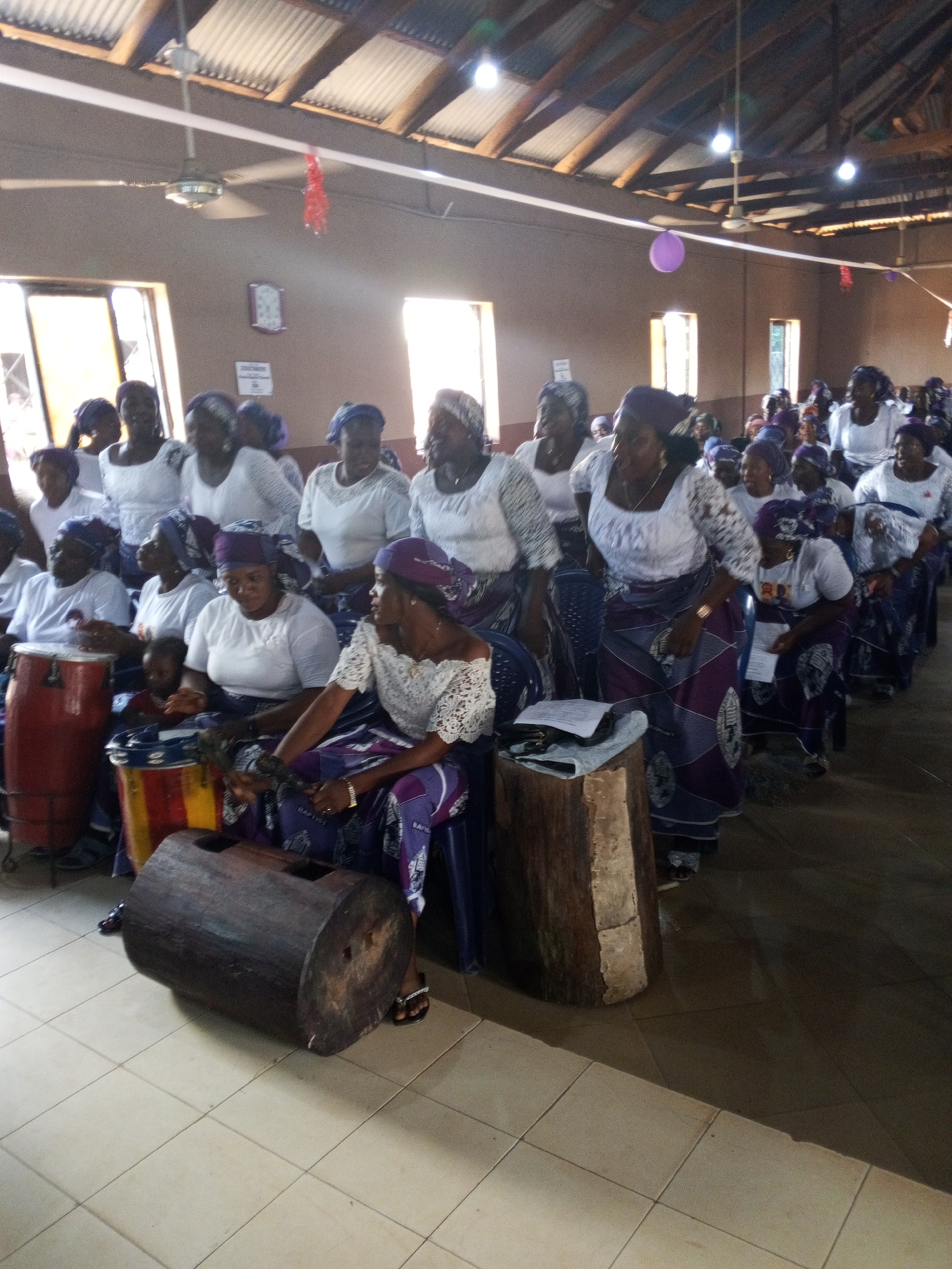 A medium shot depicts a large group of African women gathered inside a building that appears to be a church, likely in Unknown, Unknown. Most of the women are dressed in white tops and purple and white patterned skirts or wraps, with matching head coverings. They are seated in rows, some on chairs, others directly on the floor. Several women are playing musical instruments, including drums and a large wooden log-shaped instrument. The room is brightly lit by natural light coming through windows. The walls are a light brown color, and the ceiling is made of corrugated metal sheets. There are ceiling fans visible in the room. Overall, the scene suggests a cultural or religious gathering with musical performances.