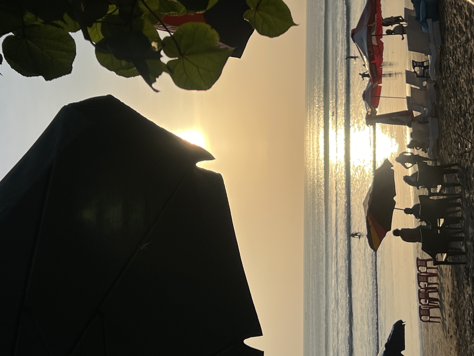 This is a photograph taken at Kuta Beach in Bali, Indonesia, during sunset. The sun is low on the horizon, casting a warm, golden glow over the scene and creating a shimmering reflection on the calm ocean waves.
In the foreground, there are several beach umbrellas and lounge chairs set up on the sand. The umbrellas are of various colors, including red and white, and a large dark umbrella is prominent in the upper left corner, casting a shadow. A few people are scattered across the beach, some sitting in chairs and others standing or walking.
In the middle ground, the water's edge is visible with gentle waves lapping onto the shore. A few individuals are in the shallow water, including a person standing on a surfboard. Further out in the ocean, there are distant figures and possibly a small boat on the horizon.
The sky is a soft gradient of pale yellow and light blue, characteristic of a sunset. Lush green leaves are silhouetted in the upper right corner, framing the scene and suggesting the presence of trees or foliage near the beach. The overall atmosphere is peaceful and serene, with the natural beauty of the sunset and the ocean dominating the view.