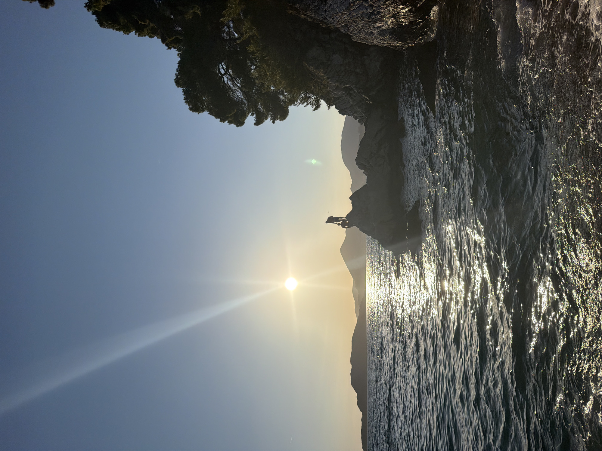 This image captures a coastal scene in Sveti Stefan, Montenegro, during golden hour. A bright sun, positioned low in the sky, emits prominent light rays and generates an intense glare that reflects across the surface of the dark blue, choppy sea, forming a shimmering path towards the viewer. The sky transitions from a clear pale blue at the zenith to a warm, golden-orange hue near the horizon.
In the mid-ground, two human figures are silhouetted against the bright water and sky, standing on a dark, jagged rock formation protruding from the sea. One figure is upright, while the other appears to be in a lower, possibly seated or crouching position. To the right, a steep, dark rocky cliff rises, densely covered with dark green coniferous trees. In the distance, multiple layers of mountains appear as dark silhouettes along the horizon. The strong backlighting creates deep shadows for all foreground elements, highlighting their forms against the illuminated sky and water.