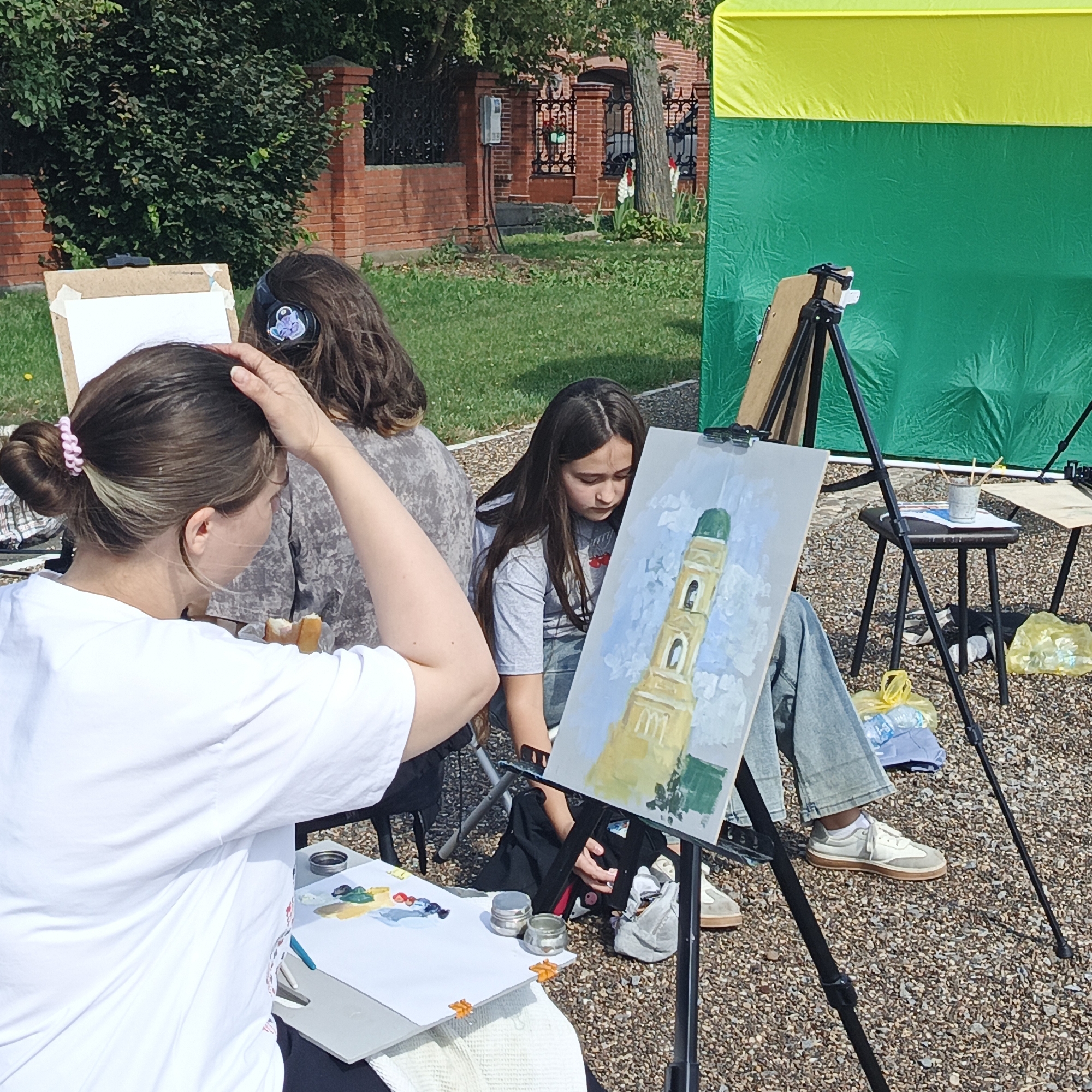 The image depicts an outdoor art class or workshop in what appears to be a park or public space with gravel ground and grassy areas. In the background, a brick fence with decorative wrought iron elements and a tree can be seen. A large green and yellow tarp or tent is also present in the background, likely providing shade.
In the foreground, three individuals are engaged in painting. The person closest to the viewer, seen from the back, is a female with her hair in a bun, wearing a white t-shirt. She is touching her head and appears to be looking at her painting setup, which includes a palette with paints. To her left, another person, likely a female, is partially visible, wearing headphones with a graphic on them. The third person, a young female with long dark hair, is seated and focused on her canvas, which features a painting of a tall, light-colored building with a green dome, possibly a bell tower or church. She is wearing a grey t-shirt and blue jeans.
Several easels are set up, supporting the canvases. A small stool with art supplies, including brushes in a container, is placed near one of the easels. Plastic bags, one yellow and one containing a bottle of water, are visible on the ground. The overall impression is that of a serene and creative outdoor activity.
The location in the city of Nikolo-Beryozovka, Russia, suggests that the building being painted might be a local landmark. The presence of the brick fence and the style of architecture visible in the background could be consistent with the historical buildings found in such a region. The natural elements of grass, trees, and the overcast sky also contribute to the environment.