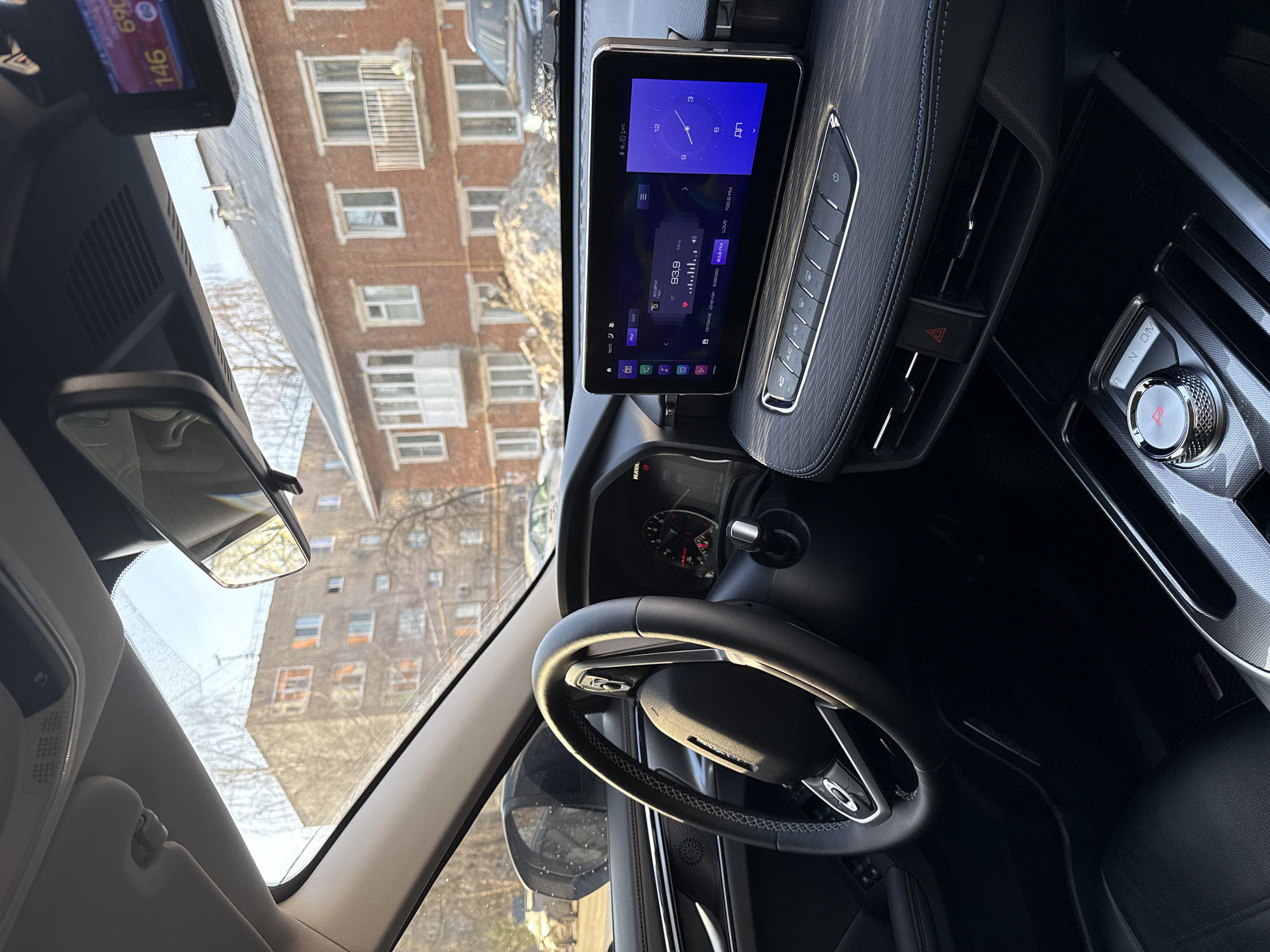 The image shows the interior of a car, specifically the dashboard, steering wheel, and center console. The infotainment system displays a clock and a radio frequency of 93.9 MHz. Other details include the rearview mirror, a dashboard camera, and control buttons for climate and car settings. The view through the windshield shows a multi-story brick building, suggesting an urban setting. Based on the radio frequency 93.9 MHz and the city names shown on the display, the image may be from a car in Izhevsk, Russia.