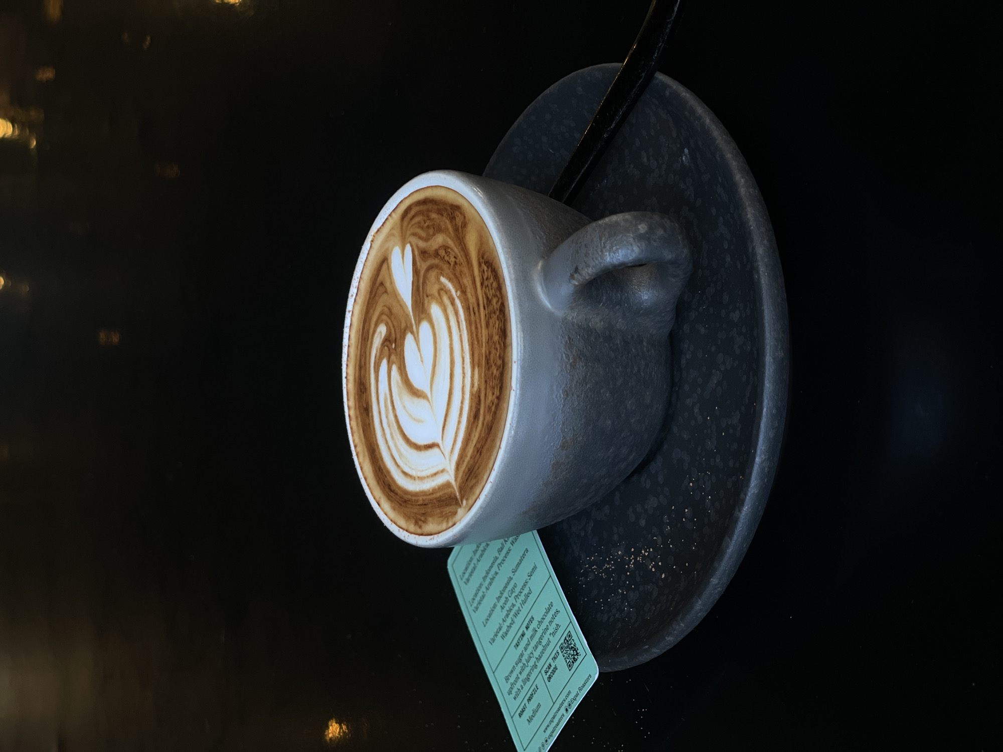 The image captures a close-up, high-angle shot of a meticulously prepared latte in a grey ceramic cup, resting on a matching saucer. The coffee features intricate latte art, consisting of several layered heart shapes or rosettas, showcasing a blend of rich brown espresso and creamy white milk. The cup and saucer have a textured, matte finish and are placed on a dark, highly reflective surface, likely a tabletop, which creates a deep, almost black background. Blurred golden and orange lights are visible as reflections in the dark surface, suggesting an indoor setting with ambient lighting.
A small, light green rectangular tag or label hangs from the cup, displaying detailed information about the coffee. The visible text on the label, according to OCR, includes "Location: Indonesia, Bali Ka", "Varietal: Arabica, Process Washed/Wet Hulled", and further "Location: Indonesia, Sumatera, Aceh Gayo". It also lists "TASTING NOTES: Brown sugar and milk chocolate upfront with juicy tangerine notes, with a lingering hazelnut 'nish." and "ROAST PROFILE: Medium". Below this, there's a QR code with the prompt "SCAN THIS QRCODE" and contact information: "www.expatroasters.com" and "@expatroasters Expat Roasters". The overall scene suggests a high-quality coffee experience, likely from a specialty coffee shop in Kuta, Indonesia.