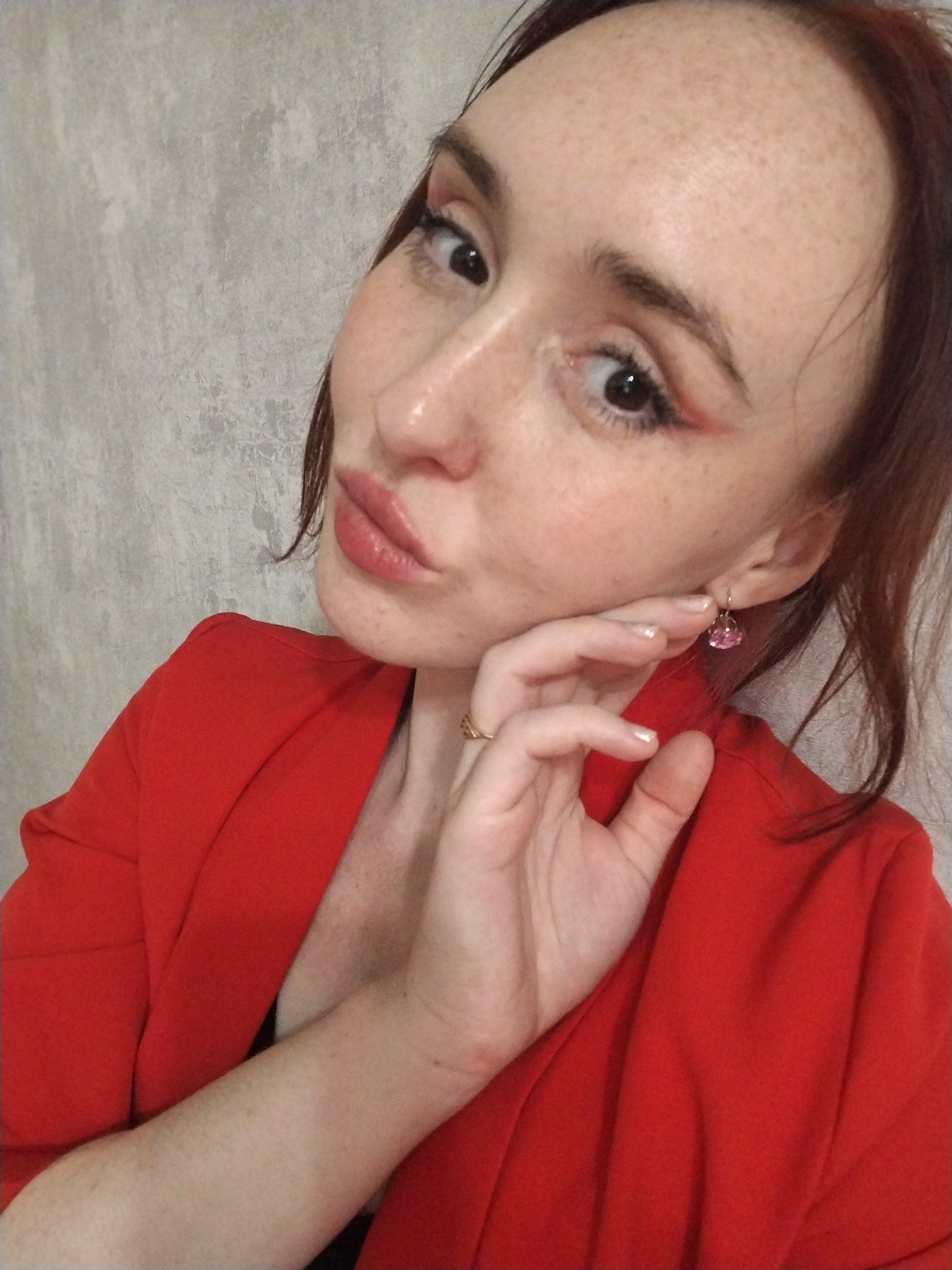 A woman with freckles, reddish-brown hair, and wearing a red blazer is posing for a selfie indoors. She has on makeup and a pink earring and her hand is near her face. The background is a grey wall. There is no information present in the image that indicates this photo was taken in Izhevsk, Russia.