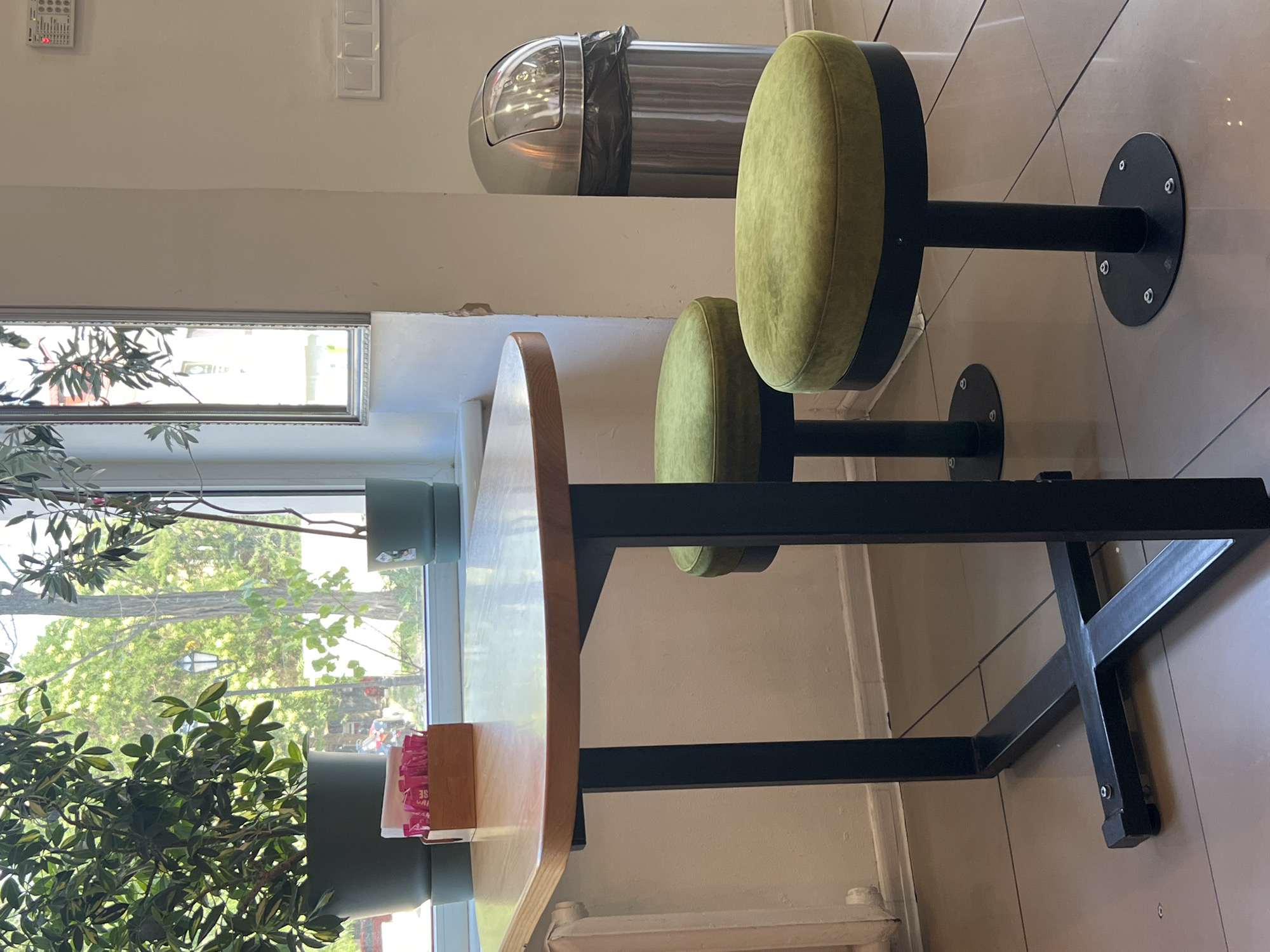 The image depicts an indoor seating area, likely a cafe or restaurant, situated in Odesa, Ukraine. In the foreground, a light-colored, oval-shaped table with a dark metal base stands prominently. Two green, plush-topped stools with dark metal stems and circular bases are placed near the table. To the left of the table, a large potted plant with green leaves is visible. A small wooden box filled with pink sachets, possibly sugar or sweetener packets, sits on the table. Behind the table and stools, a large window offers a view of trees and a street lamp outside, suggesting a daytime scene. Further to the right, a silver trash can with a domed lid is positioned against a light-colored wall. The floor is tiled with a neutral-colored, glossy finish. The overall environment appears clean and well-maintained.