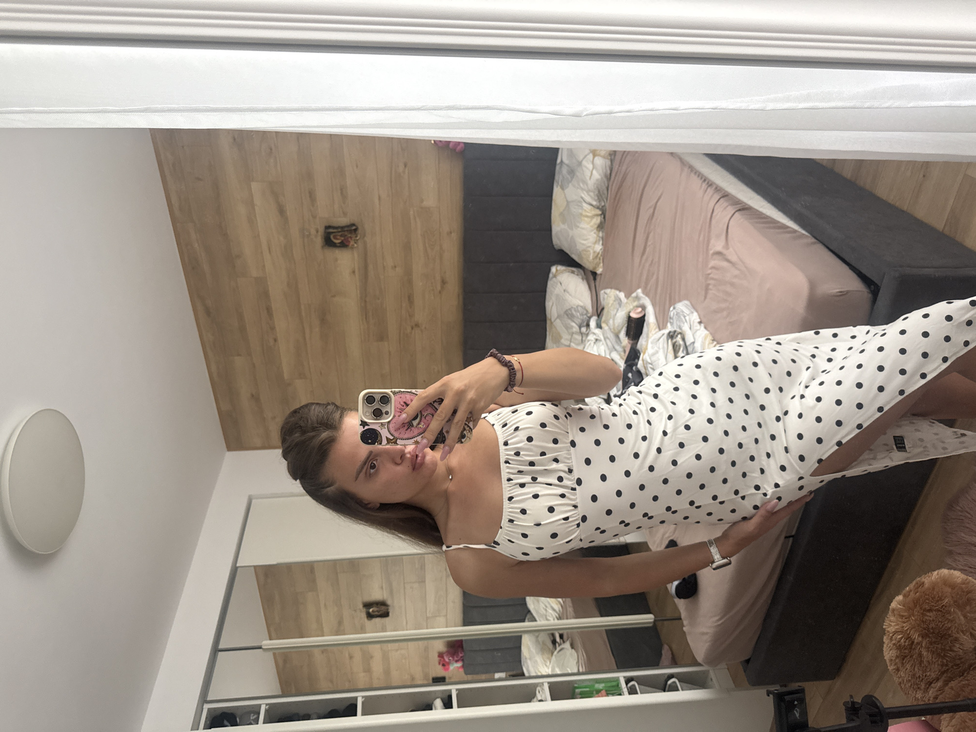 The image, which is displayed horizontally, is a mirror selfie featuring a young woman standing indoors, likely in a bedroom. She is positioned towards the center-right of the frame, facing the mirror and holding a smartphone with a pink and purple patterned case in her right hand. Her right wrist is adorned with a dark beaded bracelet, while her left wrist sports a silver Apple Watch. She has brown hair, partially styled or pulled back, and looks directly at her reflection with a neutral expression.
She is wearing a white midi-length dress with black polka dots, featuring delicate spaghetti straps and a ruched detail across the bust. The dress has a side slit near the hem, revealing a glimpse of her leg.
The background, visible as a reflection in the mirror, shows a room furnished with a bed on the right side, featuring a dark gray upholstered frame and light beige bedding with patterned pillows. The floor is made of light wooden laminate. Towards the bottom of the mirror, a white storage unit with multiple open compartments, containing various items including shoes, is visible. A circular white wall light is mounted on a plain white wall to the left of the woman. A small, dark decorative object rests on the wooden floor against the back wall, and a fluffy brown object, possibly a rug or soft toy, is on the floor next to the bed. White crown molding or trim is visible at the top edge of the reflected wall. The lighting suggests it is daytime.