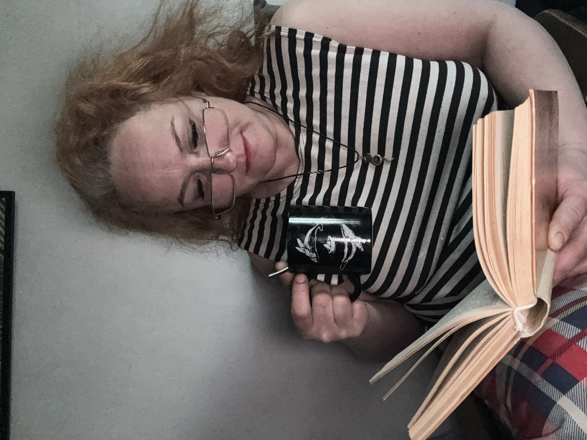 An indoor portrait shows a woman in a relaxed pose, rotated 90 degrees counter-clockwise. She has red/auburn, somewhat disheveled hair and wears wire-rimmed glasses. Her lips are slightly parted, and she looks downwards. She is dressed in a black and white striped top and wears a black cord necklace with a small metallic snake-like pendant.
In her left hand, she holds a black mug with a white graphic design featuring two stylized figures, possibly archers or mythological beings, facing away from each other with arrows drawn. Her right hand holds an open book with numerous light-colored pages and a soft, light-toned cover, suggesting it is well-read or paperback. Part of a red and blue plaid fabric, likely a blanket or clothing, is visible below the book. The background is a plain, light-colored wall, indicating an indoor setting. The image captures a moment of quiet leisure, possibly reading and enjoying a beverage in Chaikovsky, Russia.