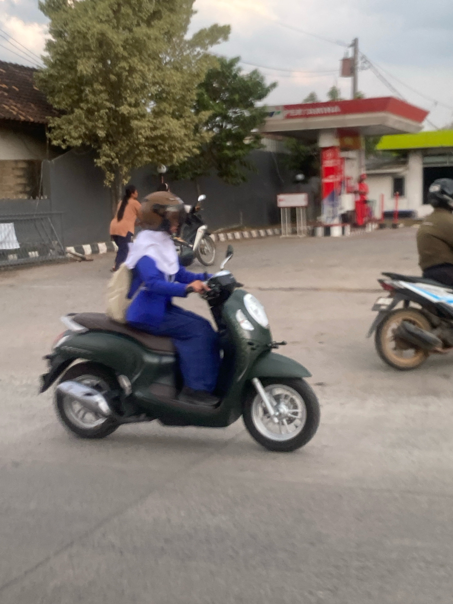 This is a medium-angle, outdoor, slightly blurred photograph taken during the day. The scene appears to be on a paved road in front of a gas station. In the foreground, a person wearing a blue uniform and a white hijab is riding a dark green scooter. The person is wearing a helmet and a backpack. The scooter's wheels are clearly visible, and the rear tire is in focus. In the mid-ground, another person in an orange top and dark pants is walking away from the camera. A white scooter is parked behind them. Behind the people, there is a red and white gas station canopy with visible signage. To the left of the gas station, there is a building with a tiled roof and a dark fence. Further to the left, a large tree with light green foliage is visible. The sky is overcast. The overall environment suggests a typical urban setting in Indonesia, likely in the city of Metro, given the visual cues of the architecture and the presence of a gas station with Indonesian branding.
