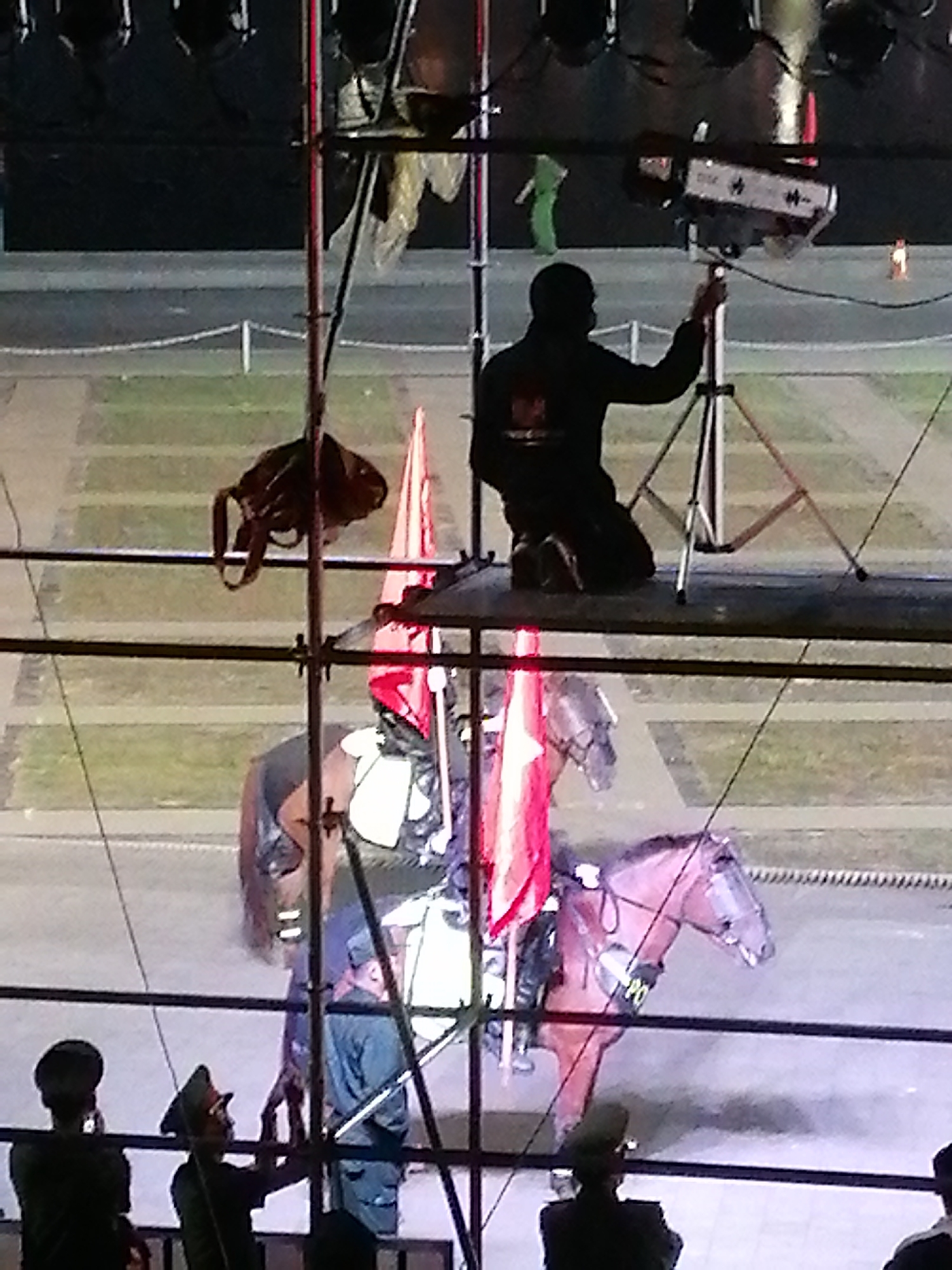 The image depicts an outdoor event, possibly a parade or ceremonial gathering, with a focus on a horse and rider. In the foreground, several individuals in uniform, possibly military or police, are visible. One person in the center appears to be interacting with a stand or mechanism, while another to the left is holding a phone or similar device. Behind them, a horse is the central subject, adorned with some sort of decorative covering or armor. A rider is mounted on the horse, also wearing a uniform and holding a red flag. Two other red flags are visible near the horse and rider. In the background, a person in dark clothing is operating a lighting fixture on a tripod, and further back, another person in green is visible. The setting appears to be a paved area with grassy sections and some fencing, suggesting an open space within a city. The overall impression is one of a staged event with a ceremonial element. The specific location in Da Lat, Vietnam cannot be determined from the image alone.
