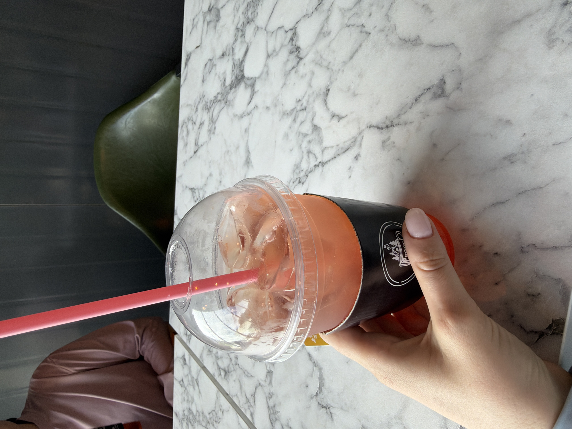 A close-up shot shows a hand holding a clear plastic cup filled with a pink, ice-laden beverage. A pink straw extends from the cup. The cup is encased in a black cardboard sleeve with a white logo featuring trees and a mug. The cup rests on a white marble tabletop. In the blurred background, a person in a pink shirt is visible, and a dark green chair is placed against a dark gray wall. The scene appears to be an indoor establishment, possibly a cafe or restaurant, in Seo-myeon, South Korea.