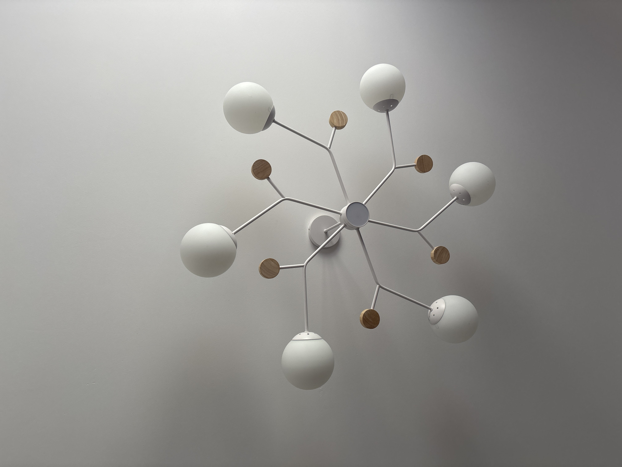 A low-angle shot depicts a modern light fixture mounted on a gray wall. The fixture features a central hub with multiple white branches extending outwards. Each branch supports a round, white light globe. Additionally, small, flat, wooden discs are attached to some of the branches. The setting appears to be an interior space, likely a room with a neutral color scheme. The location is not discernible from the image.