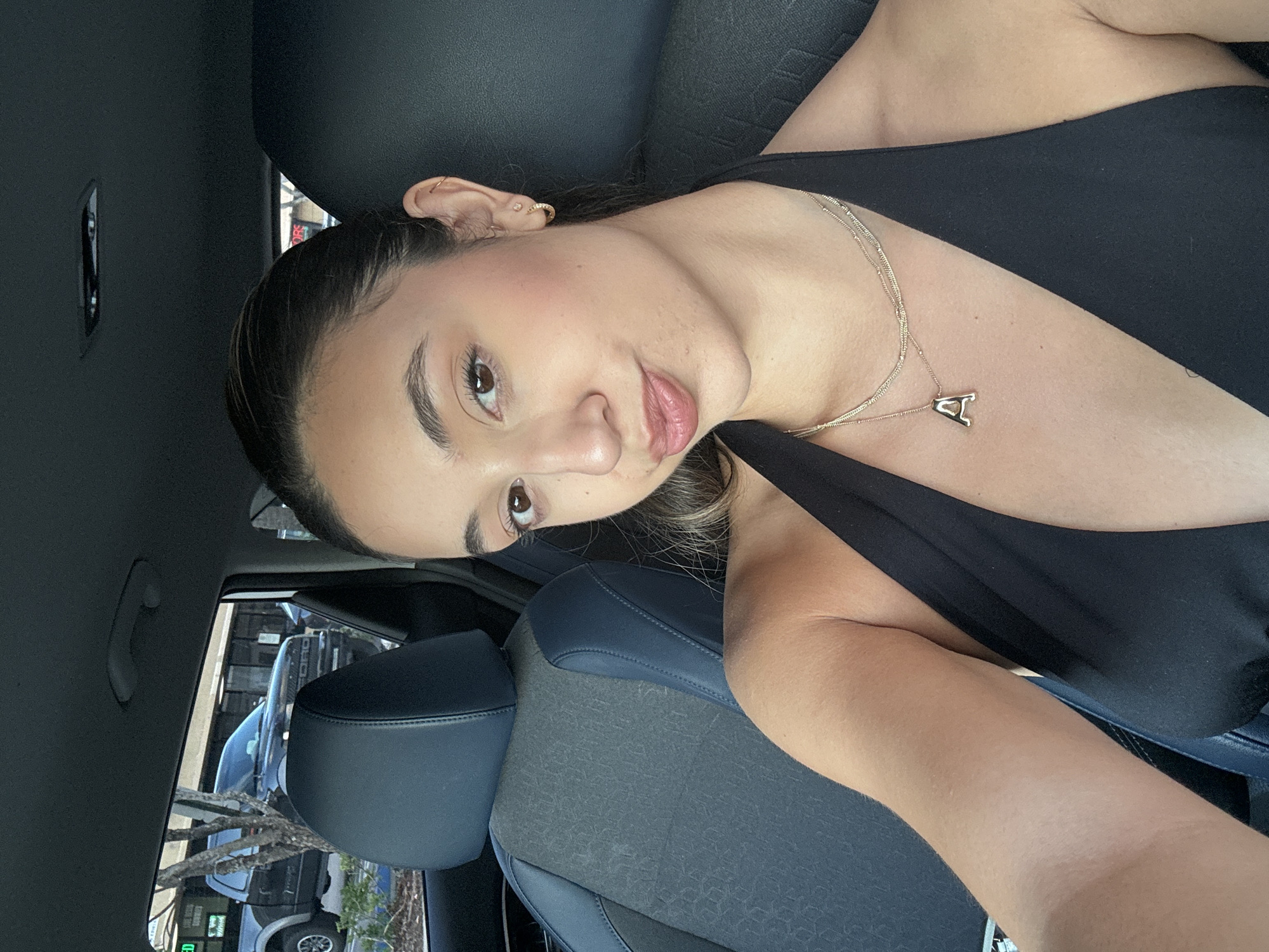 A female with dark hair pulled back, wearing a black halter-neck top, is seated in the front passenger seat of a vehicle, taking a selfie. She wears multiple small gold-colored earrings in her left ear and two gold-colored necklaces, one with an "A" pendant. The car's interior features dark gray fabric seats with dark blue leather trim. Visible through the window are outdoor commercial buildings, a dark-colored Ford pickup truck, and various trees and shrubs. A partially visible red sign displaying "ORS" is discernible in the background. The scene is located in the city of Deerfield Beach, United States of America.