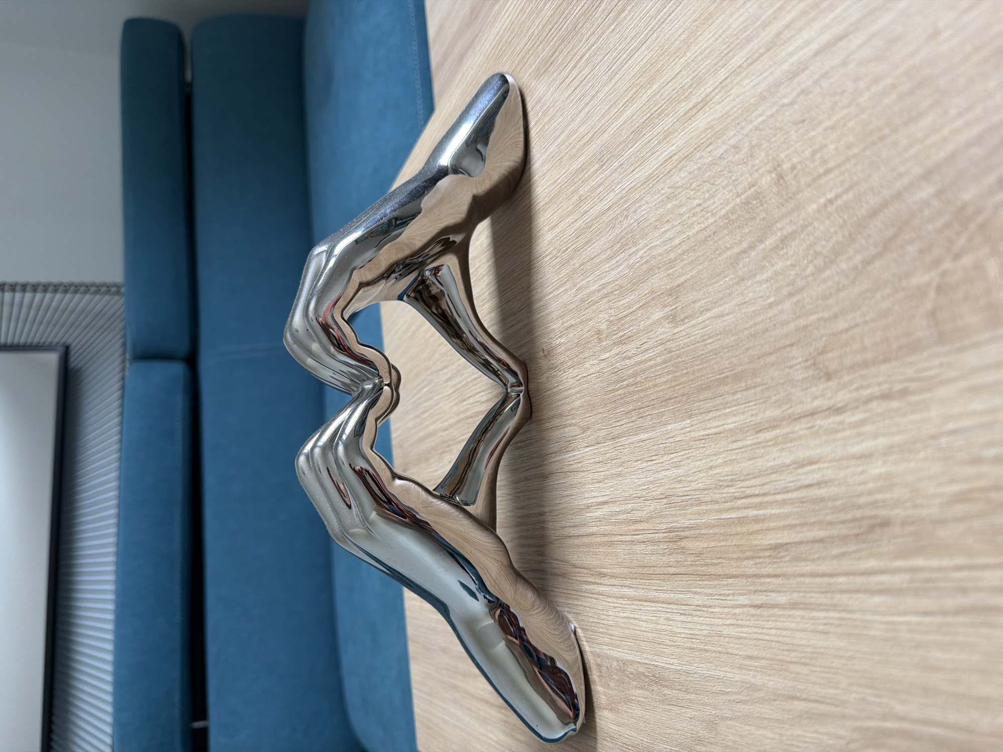 The image shows a close-up, eye-level shot of a polished chrome sculpture of two human legs intertwined, resting on a light wood-grain surface. The sculpture is abstract and stylized, with smooth, flowing lines that reflect the surrounding environment. The legs appear to be in a dynamic pose, suggesting movement or dance.
To the left of the sculpture, out of focus, are two segments of a bright blue upholstered surface, possibly a sofa or seating. A portion of a grey, ridged surface and a sliver of a white wall are also visible in the far left background. The lighting is bright and casts subtle reflections on the chrome sculpture, indicating an indoor setting with ample light, possibly natural light from a window or artificial lighting. The overall mood is sleek and modern, with a focus on form and texture. There is no visible text in the image.