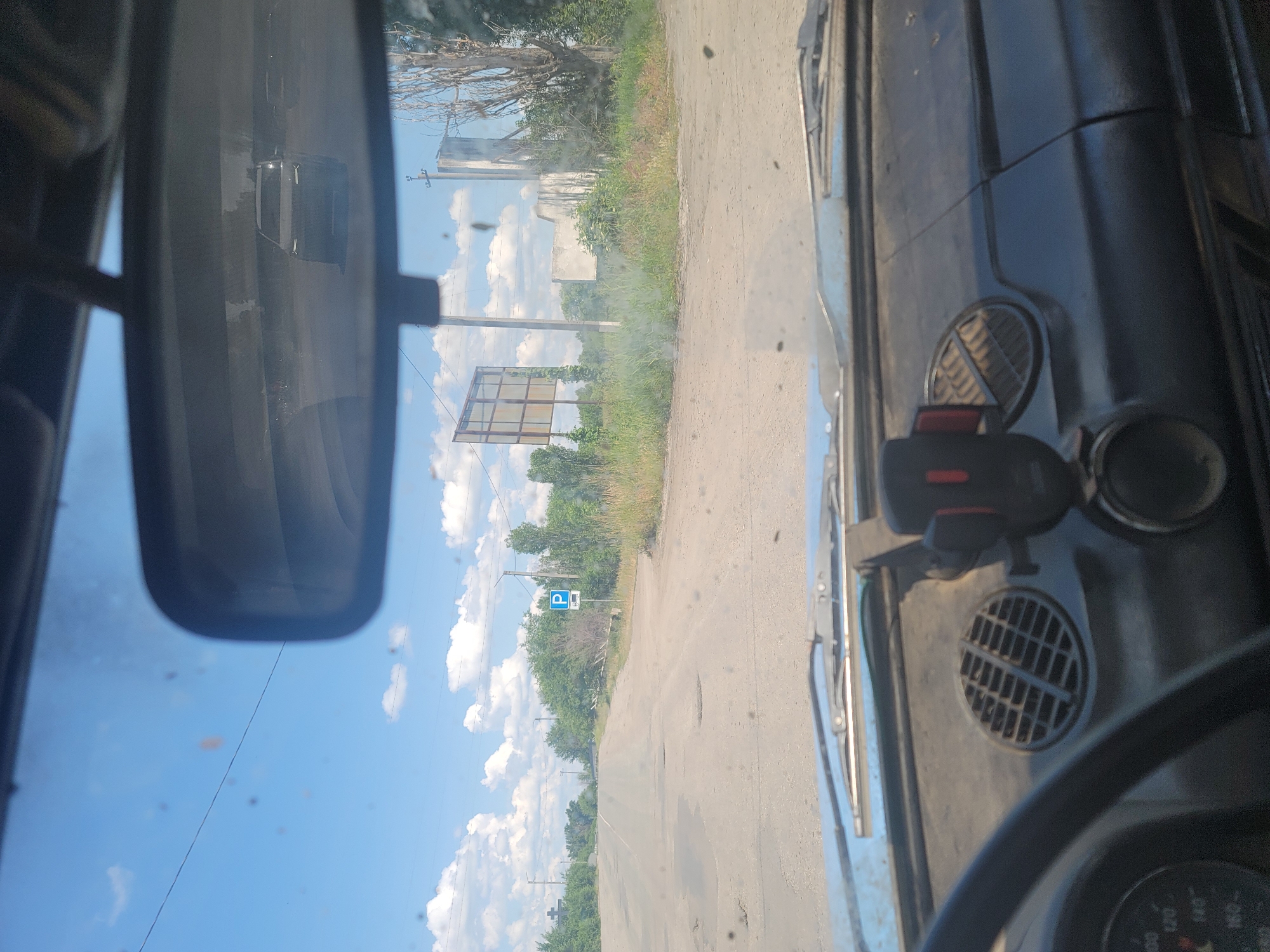 A view from inside a car looking out through the windshield, showing a road with potholes. A parking sign is visible on the right side of the road. In the rearview mirror, a white car is reflected. The sky is blue with scattered clouds. The dashboard of the car is partially visible in the foreground. The scene appears to be in Pereshchepyne, Ukraine.