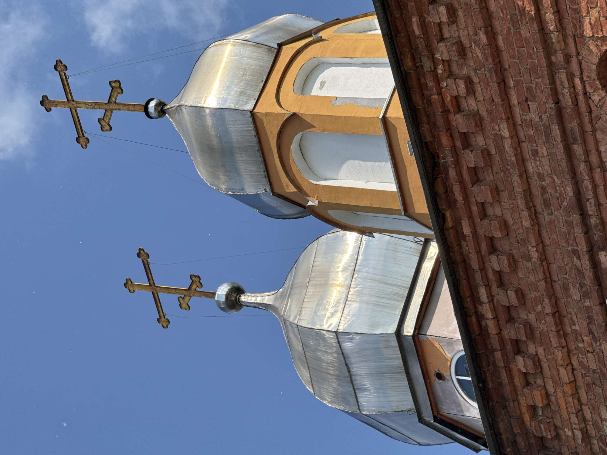 The image shows the domes of an Orthodox church in Chudovo, Russia, against a bright blue sky with a few scattered clouds. The foreground is dominated by a brick wall with decorative elements. Two metallic domes, one with a golden cross and another with a similar cross, rise above the wall. The domes are topped with multi-barred crosses, typical of Eastern Orthodox Christianity. The dome on the right is attached to an orange structure with arched windows. The overall scene suggests the exterior of a religious building.