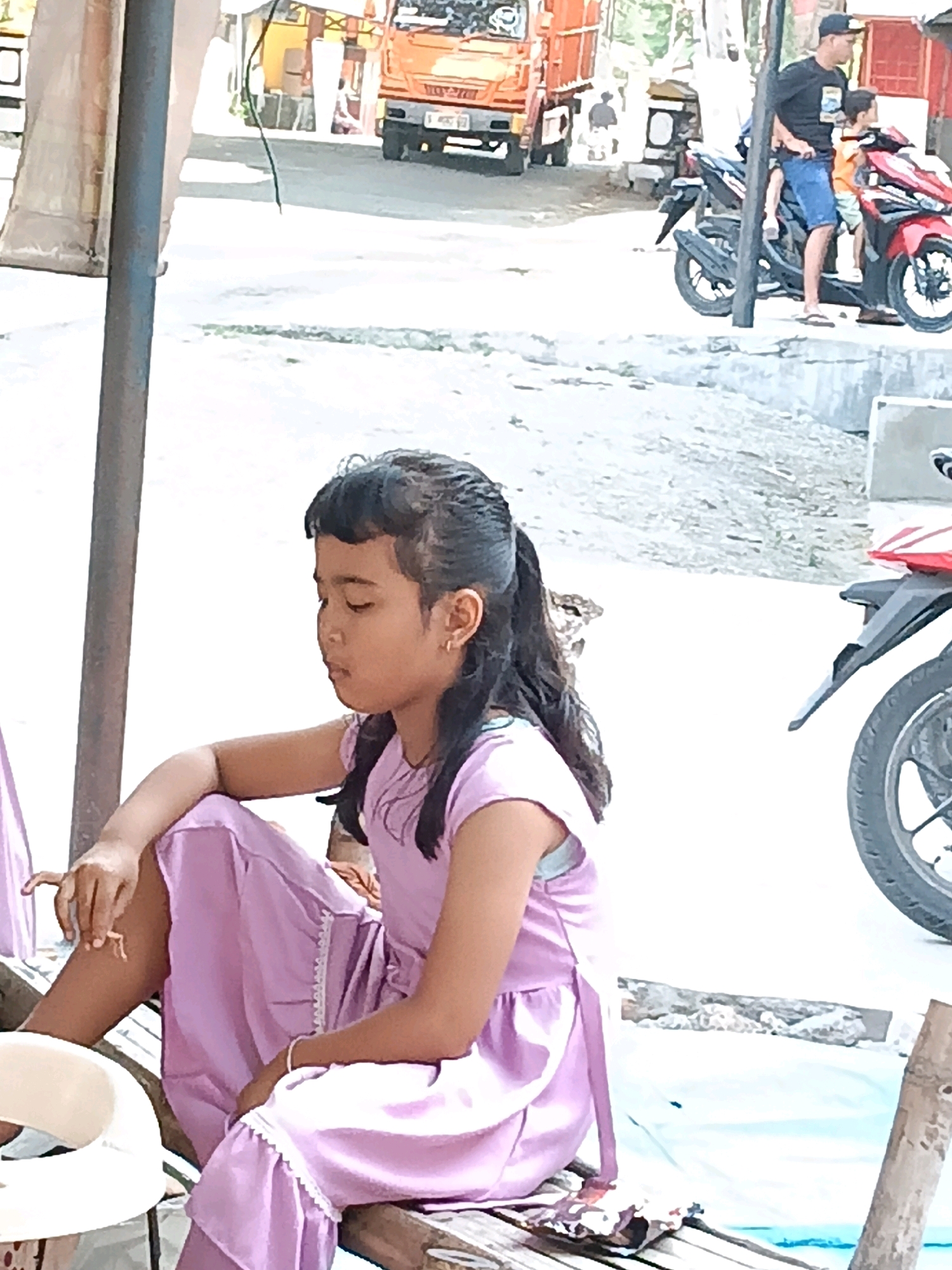 A young girl with dark hair pulled back in pigtails sits on a wooden structure, wearing a light purple dress with lace trim. She is looking down, and her hands are resting on her lap. In the background, a man wearing a black t-shirt, blue shorts, and a baseball cap stands next to a red motorcycle. He is holding a younger child, who is wearing an orange shirt. A large orange truck is parked behind them. The scene appears to be an outdoor area, possibly a roadside or market, in Kedungtuban, Indonesia. The ground is covered in gravel and dirt.