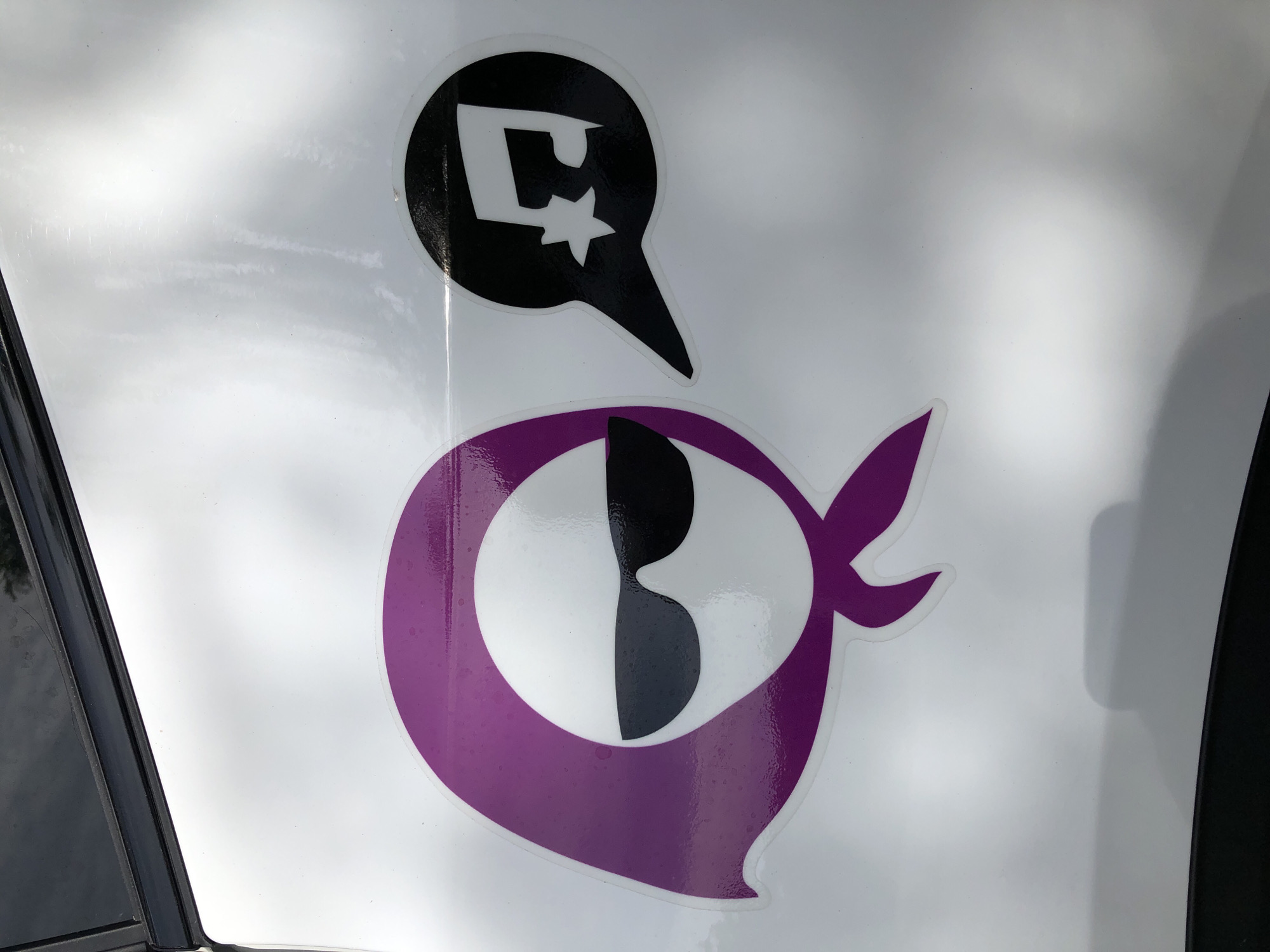 An outdoor, close-up, eye-level shot captures a portion of a white vehicle, likely a car door, featuring two stickers. On the left, a sticker depicts a stylized, white, faceless head covered by a purple headscarf, wearing black sunglasses. To the right of this figure, a black speech bubble contains a white musical note symbol and a small star. The stickers are applied to the vehicle's exterior, which appears to be in good condition, reflecting light. The background consists of the white surface of the vehicle, with a dark strip at the top, possibly the edge of a window or trim. There is no indication of a specific location within the city of Đà Nẵng, Vietnam, as the image focuses solely on the stickers on the vehicle.