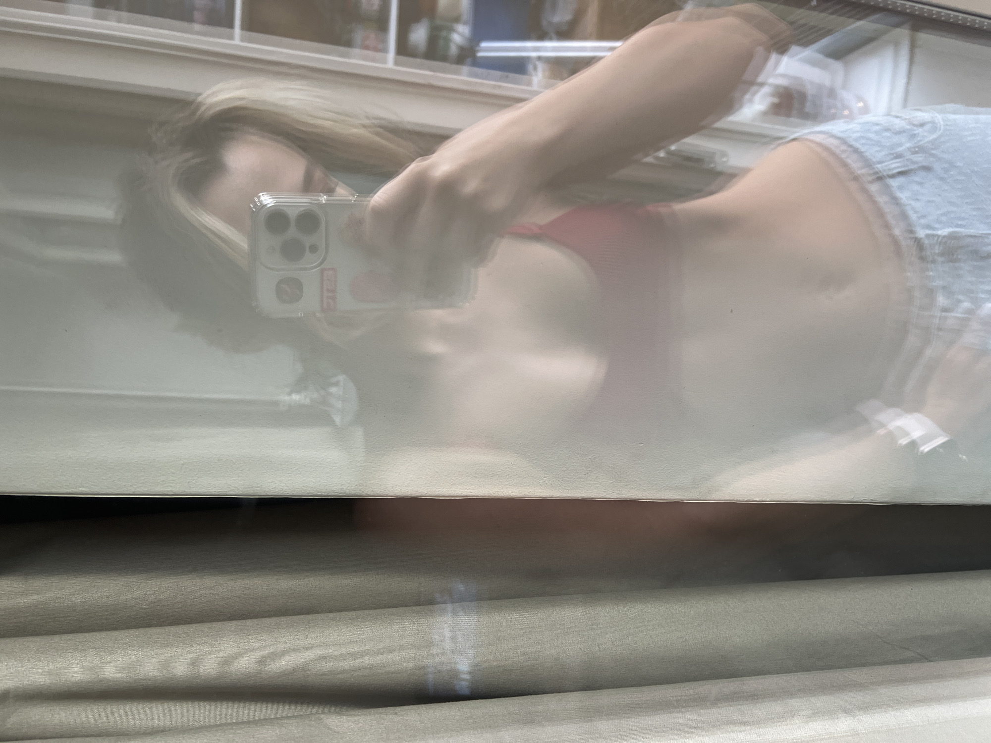 The media file displays a reflection of a person taking a selfie, likely in a mirror or a window, with the setting identified as Voronezh, Russia.
**Subjects:** The primary subject is a person, appearing to be a young woman, with long blonde hair. Her face is obscured by the smartphone she is holding. Her right arm and hand are visible, gripping the phone. Part of her torso is reflected, showing her wearing a red top (possibly a bra or bikini top) and light blue denim shorts or skirt.
**Objects:** The central object is a smartphone, specifically an iPhone with three camera lenses, housed in a clear protective case. The case has several small stickers, including a red rectangular one with faint white text (possibly "STG" or similar) and another resembling a red heart character. The background within the reflection appears to show a room with blurred objects, possibly shelves or furniture. Below the reflective surface, there are horizontal window blinds or shades, suggesting an indoor setting looking outwards or at a mirror placed in front of a window.
**Activity or event taking place:** The person is actively taking a self-portrait (selfie) using their smartphone, capturing their reflection.
**Notable details:** The image has a soft, somewhat blurry quality due to it being a reflection and possibly due to motion. Light appears to be coming from the front, illuminating the reflection. The blonde hair is spread out, adding to the soft focus. The lower portion of the image clearly shows the texture and folds of closed blinds, with a narrow gap at the top allowing some light to pass through. The overall lighting suggests daylight.
**Visible text:** Faint white text is visible on a red rectangular sticker on the smartphone case, though it is largely illegible.