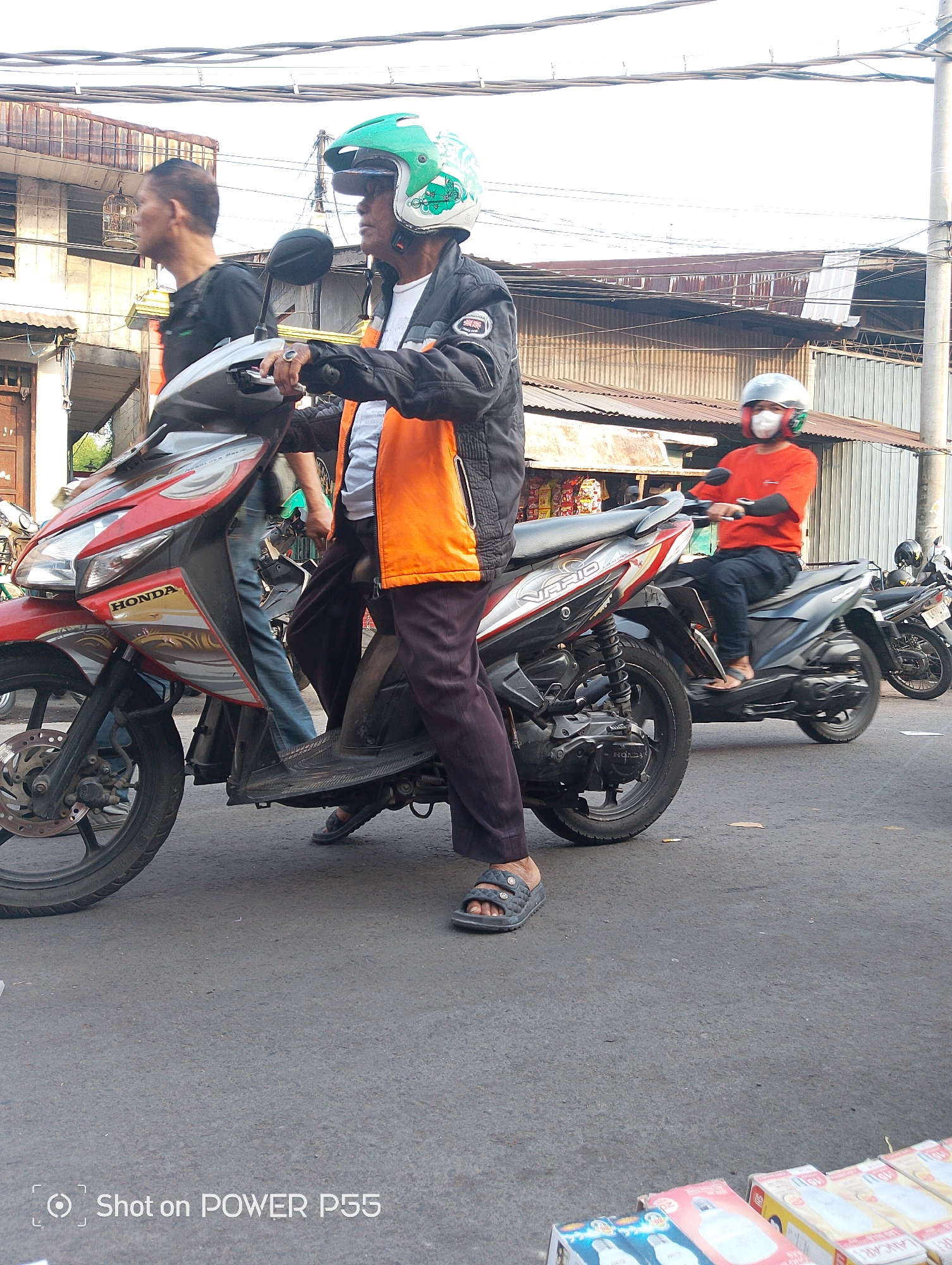 Two men and a person in an orange shirt on motorcycles are on a street in Surabaya, Indonesia. The person in the orange shirt is wearing a white mask and a silver helmet, while the man in front is wearing a green helmet. The man in front is wearing a black jacket with an orange vest and dark pants. The second man in the background is wearing a black shirt and blue jeans. The motorcycles are parked on a paved street. There are buildings in the background with corrugated metal roofs. The sky is overcast.