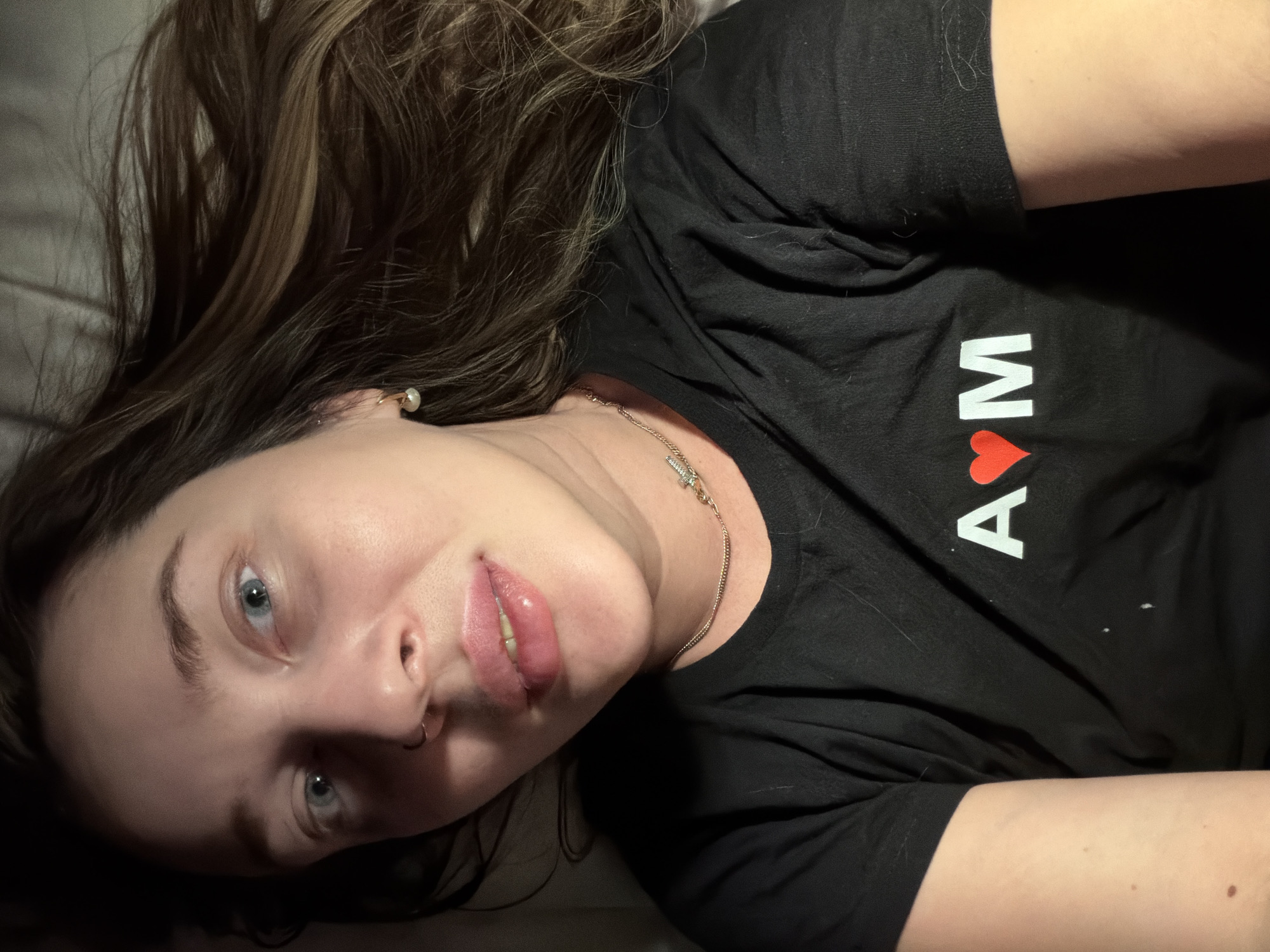 A woman with light skin and blue eyes is lying down, looking at the camera. She has brown hair and is wearing a black T-shirt with the letters "A" and "M" in white, separated by a red heart symbol. She has a nose ring and a delicate gold necklace. Her surroundings are dark and out of focus, but she appears to be on a soft surface, possibly a bed. There is no information to suggest a specific location in Moscow, Russia.