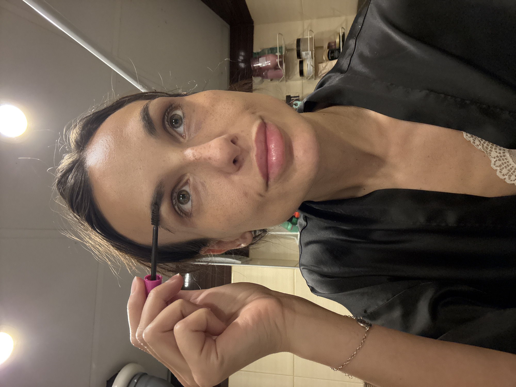 The image shows a woman applying mascara to her eyebrows in a bathroom. She is wearing a black satin robe and a white lace camisole. The bathroom appears to be in a residential building in the city of Pleven, Bulgaria. The lighting suggests an indoor setting, possibly at night or in a room with limited natural light. Shelves with toiletries are visible in the background. The woman has fair skin with freckles and is looking upwards while applying the makeup.