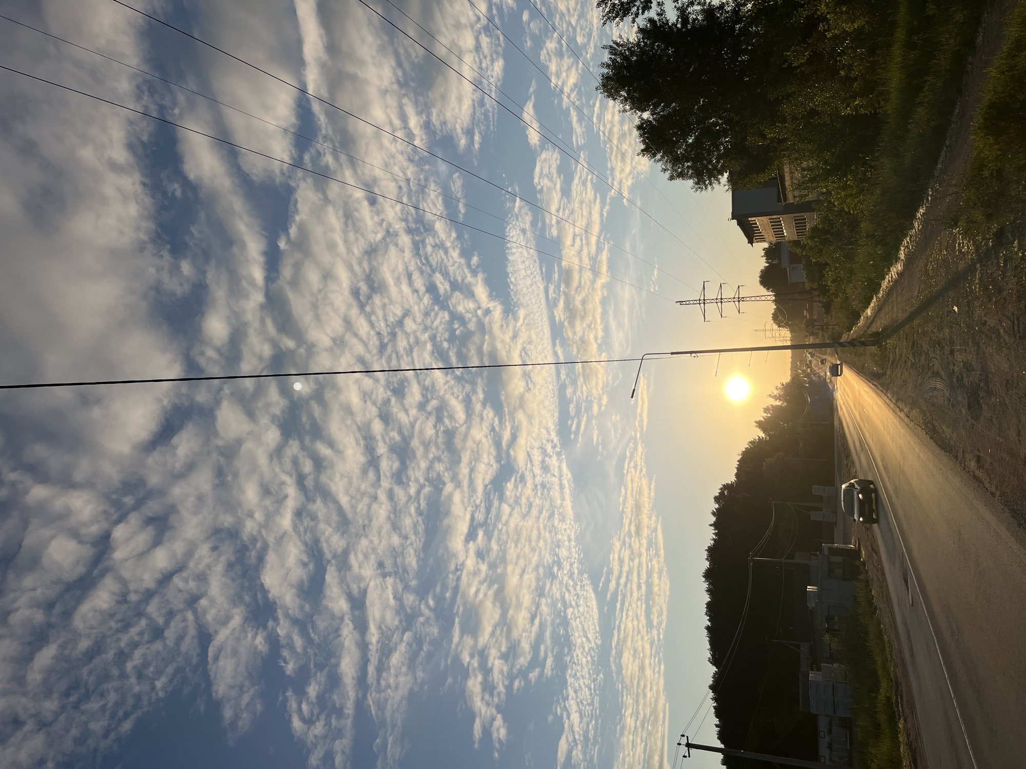 This image captures an outdoor scene in the city of Chaikovsky, Russia, during golden hour, characterized by a low sun angle and strong backlighting. The upper two-thirds of the frame is dominated by a light blue sky patterned with numerous white, altocumulus-like clouds. Overhead power lines traverse the upper sky, extending diagonally across the frame.
The sun, a brilliant white orb, is positioned low on the horizon towards the lower center-right, emitting a strong glare that creates a prominent golden reflection across the asphalt road surface. The road, appearing as a two-lane thoroughfare, recedes into the distance. A dark-colored vehicle, possibly a sedan or small SUV, is visible in the mid-ground, traveling away from the camera, its form partially silhouetted by the intense sunlight reflecting off the road. Its headlights appear to be on. A second, more distant vehicle is faintly visible further down the road.
To the left of the road, a dense line of dark green trees forms a silhouetted backdrop. Below the trees, stacks of light-colored construction blocks are visible on the ground, suggesting an ongoing or paused construction project. A utility pole with power lines is positioned near the left edge of the frame.
On the right side of the road, a dirt or gravel path runs parallel to the asphalt. This path is bordered by tall green grass and a variety of leafy trees and bushes. A street light pole stands in the immediate foreground on the right, casting a long shadow diagonally across the path. In the middle distance on the right, multiple tall metal electricity transmission towers are visible, alongside a multi-story building with numerous windows. No individuals are discernible in the scene.
