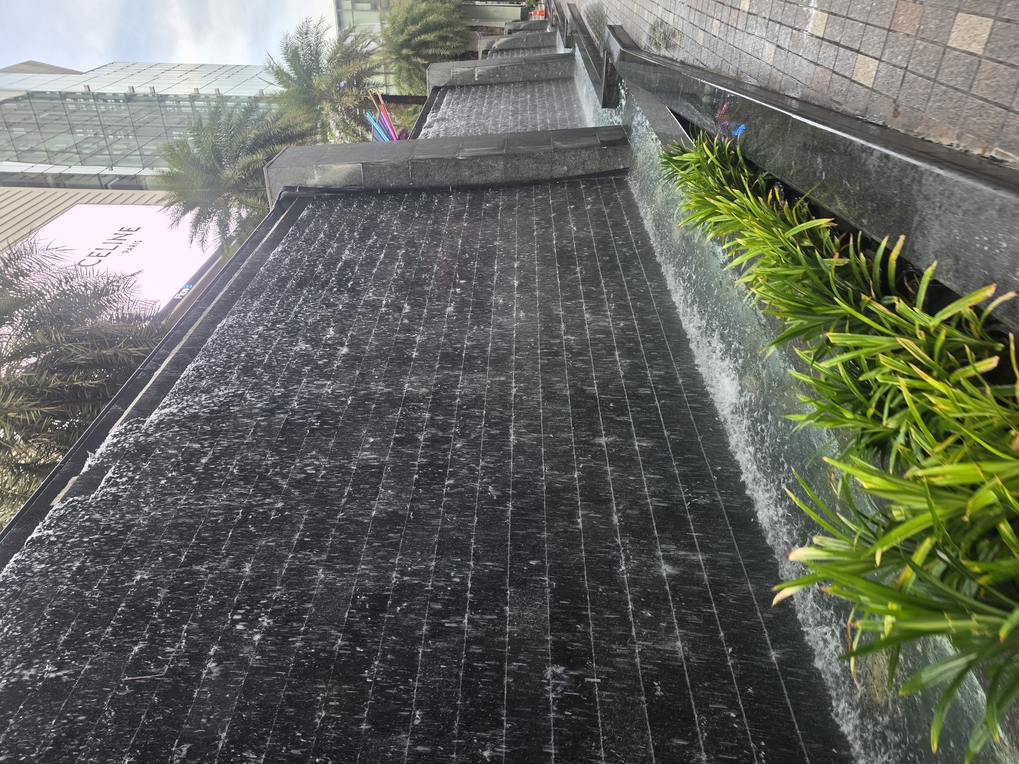 The image displays an upscale urban outdoor scene in Bangkok, Thailand, featuring a modern, multi-tiered water feature. Water cascades continuously down several dark, textured surfaces, creating a shimmering waterfall effect. To the right, a vibrant strip of lush green, spiky-leaved plants grows along the edge of the water feature, with a tiled walkway or wall beyond them.
In the background, on the left, tall palm trees are visible, characteristic of a tropical climate. Behind the water feature and trees, a modern building with a striking glass and metal facade rises. A large, prominent white sign is affixed to this building, clearly displaying the text "CELINE PARIS," indicating a luxury retail brand, likely part of a high-end shopping mall or commercial complex.
The scene is set during the daytime, under a partly cloudy sky. No people are visible, and the overall atmosphere is one of sophisticated urban tranquility.