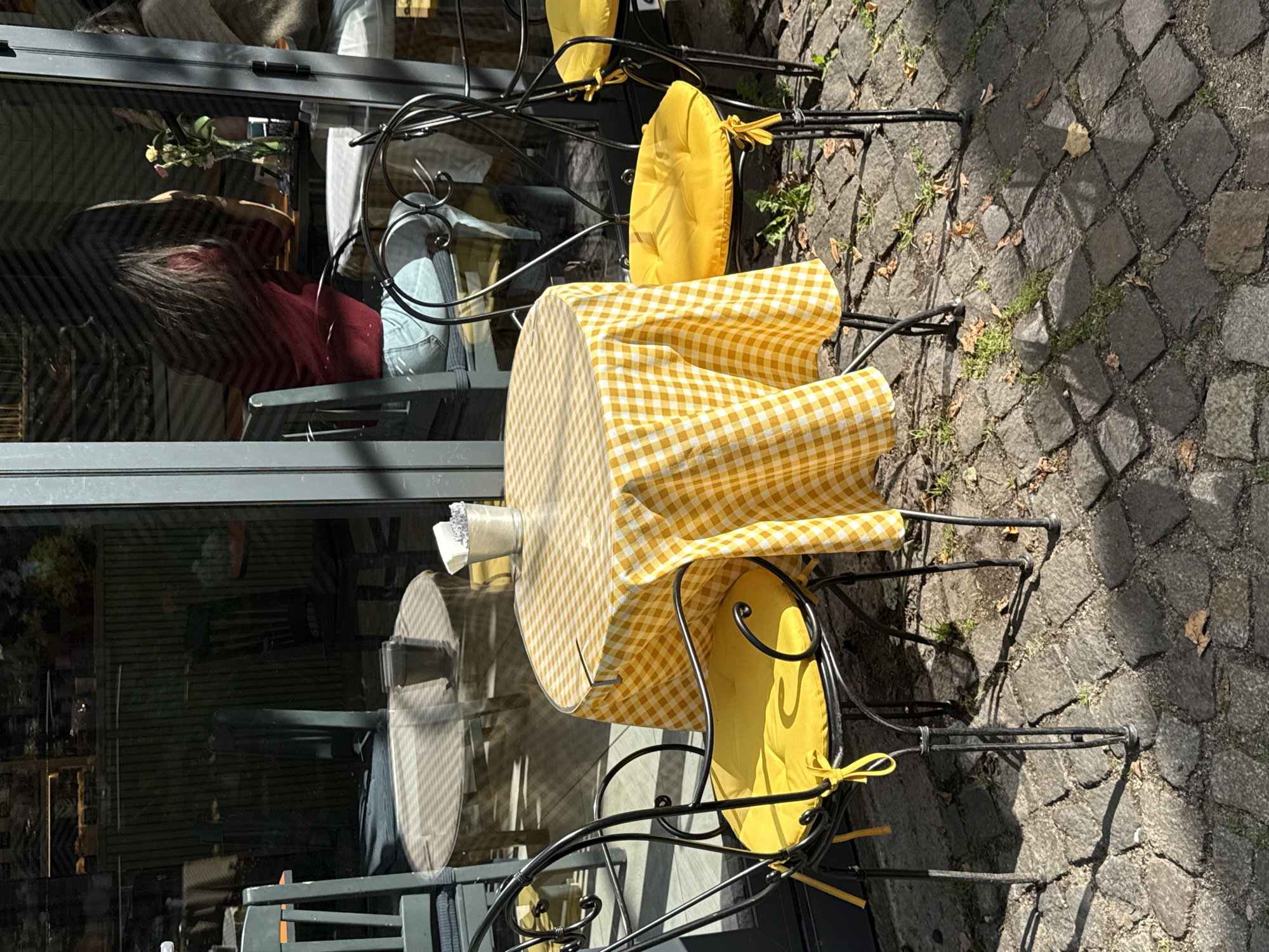The image shows an outdoor seating area of a cafe with a round table covered with a yellow and white checkered tablecloth. Two wrought-iron chairs with bright yellow cushions are placed at the table. A small metal bucket with napkins or tissues sits on the table. In the reflection of the cafe window, two women are visible inside. One woman in a red top is seated at a table. The cafe appears to be located on a cobblestone street. The scene suggests a pleasant day with sunlight casting shadows. There is no specific information in the image to determine the location as Kaliningrad, Russia.