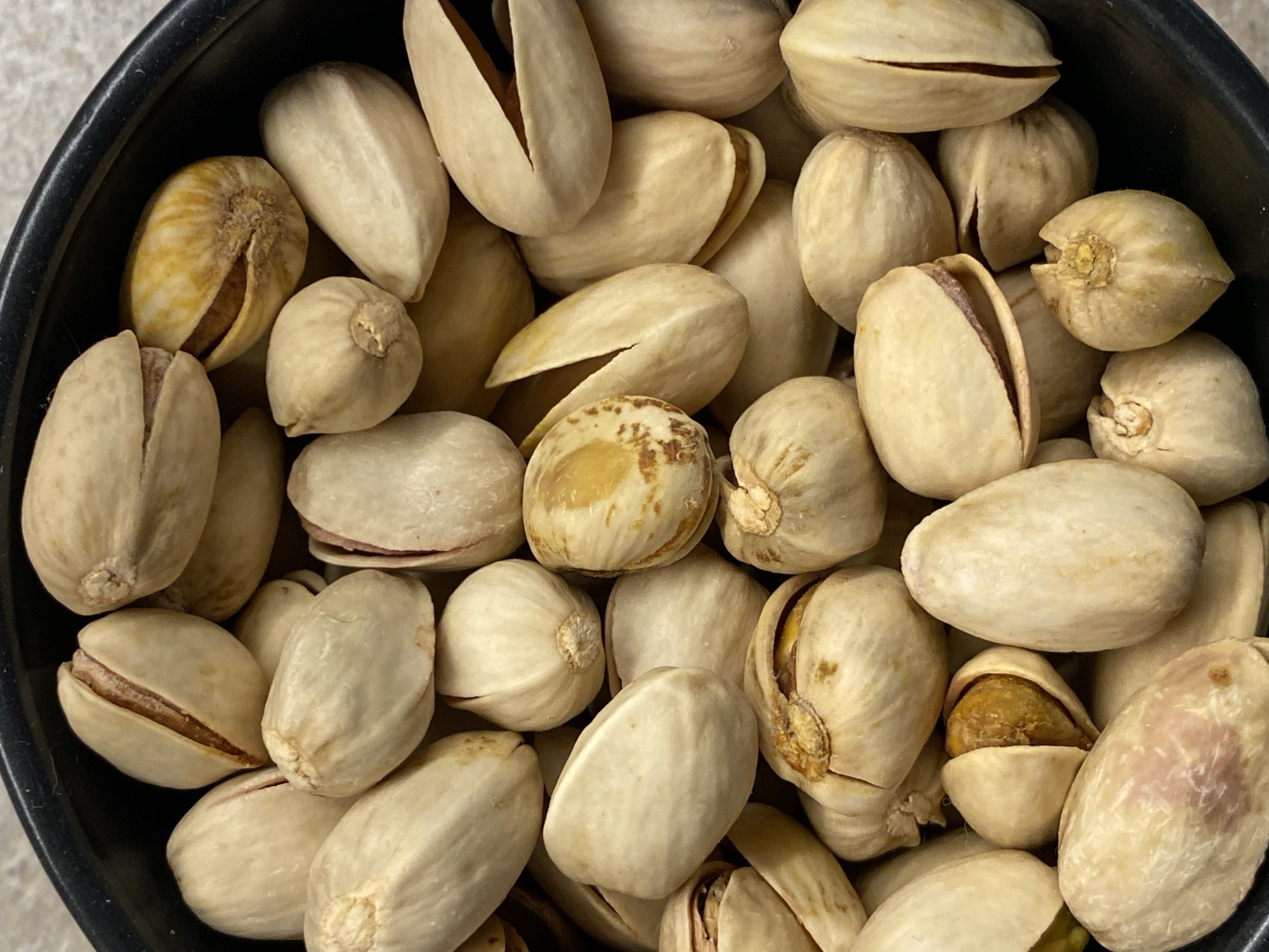 The image depicts a close-up view of a bowl filled with pistachios. The pistachios are of varying shapes and sizes, some with open shells exposing the nut inside, while others are closed. The shells are a light beige color with darker markings. The bowl appears to be black and is partially visible at the edge of the frame. The background is a light grey color. No people, specific actions, or interactions are visible in the image. The location in Budva, Montenegro is not evident from the image.