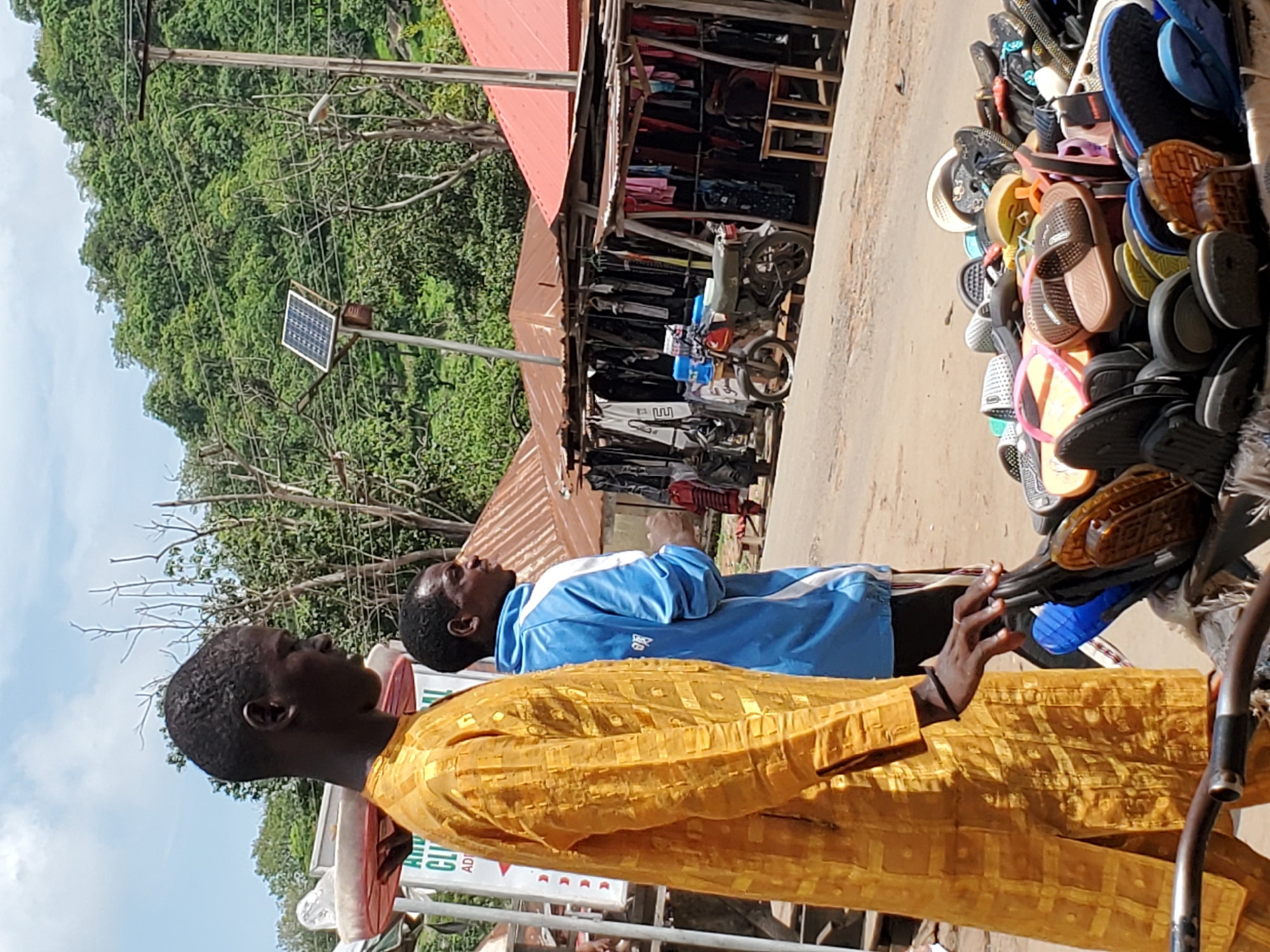 In Mararaba, Nigeria, two men stand in front of a street vendor stall displaying sandals. The man closest to the viewer wears a long-sleeved yellow shirt with a textured pattern. The second man wears a blue jacket with white accents. Behind them, the vendor stall is stocked with a variety of clothing. A motorcycle is parked within the stall area. In the background, a hillside covered with trees is visible. A solar panel is mounted on a pole near power lines.