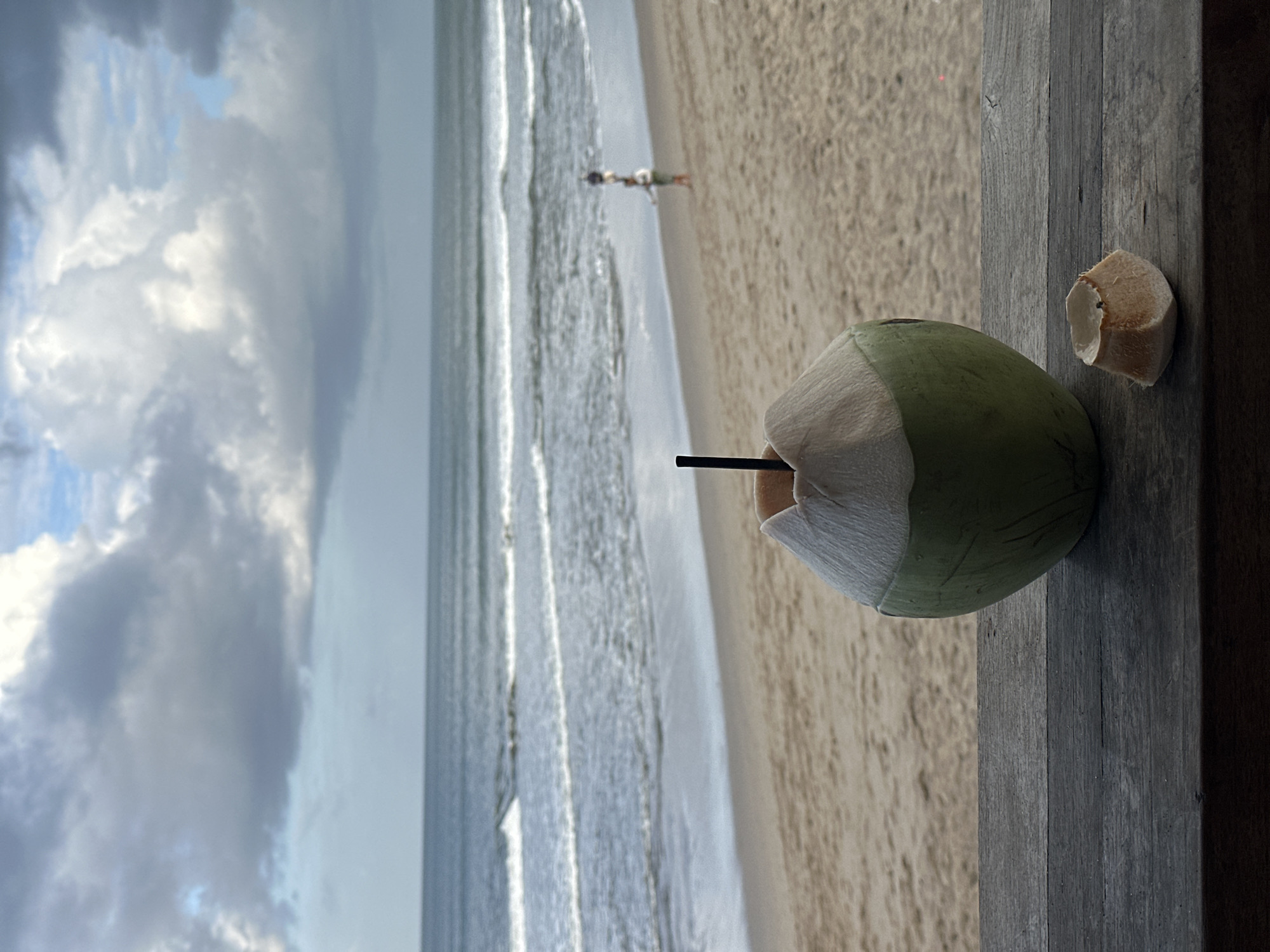 A young green coconut with a black straw sits on a weathered wooden surface in the foreground. To the right of the coconut is a small, jagged piece of coconut shell. In the background, a sandy beach meets a calm ocean with gentle waves. The sky above is filled with dramatic, gray clouds interspersed with patches of blue. Further down the beach, a person is visible walking near the water's edge. The scene appears to be a serene beach setting in North Kuta, Indonesia.