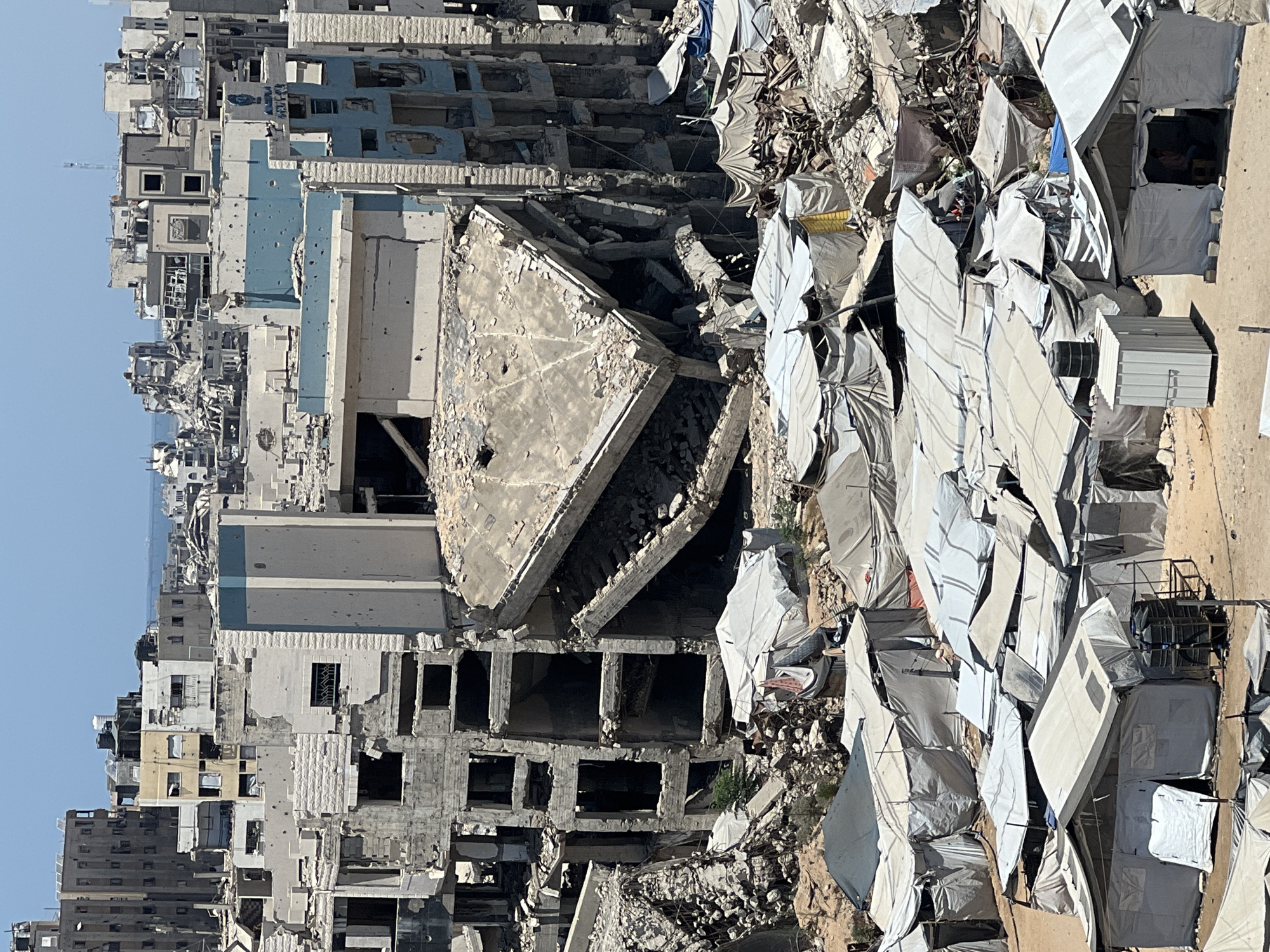 The image depicts a densely populated area within the city of Gaza, Palestinian Territories, characterized by widespread destruction and makeshift shelter. In the background, multi-story buildings stand in various states of ruin, with collapsed walls, exposed rebar, and fragmented concrete structures. Many buildings show signs of damage from shelling or bombing, with numerous bullet holes visible on their facades.
In the foreground and middle ground, a large encampment of temporary shelters has been established amidst the rubble. These shelters are primarily made of white and grey canvas tarps, arranged closely together to form a maze-like settlement. Some shelters appear to be more robust, with wooden or metal frames, while others are simple tent-like structures. Various objects are visible within and around the shelters, including water tanks, storage containers, and personal belongings. The ground is a mix of dirt, sand, and debris from the destroyed buildings.
The overall scene conveys a sense of displacement and hardship, with the destroyed urban landscape serving as a stark backdrop to the temporary housing arrangements of the displaced population. The sky is clear and bright, suggesting a sunny day. There are no visible individuals clearly engaged in any specific action, though glimpses of people can be seen inside some of the tents.