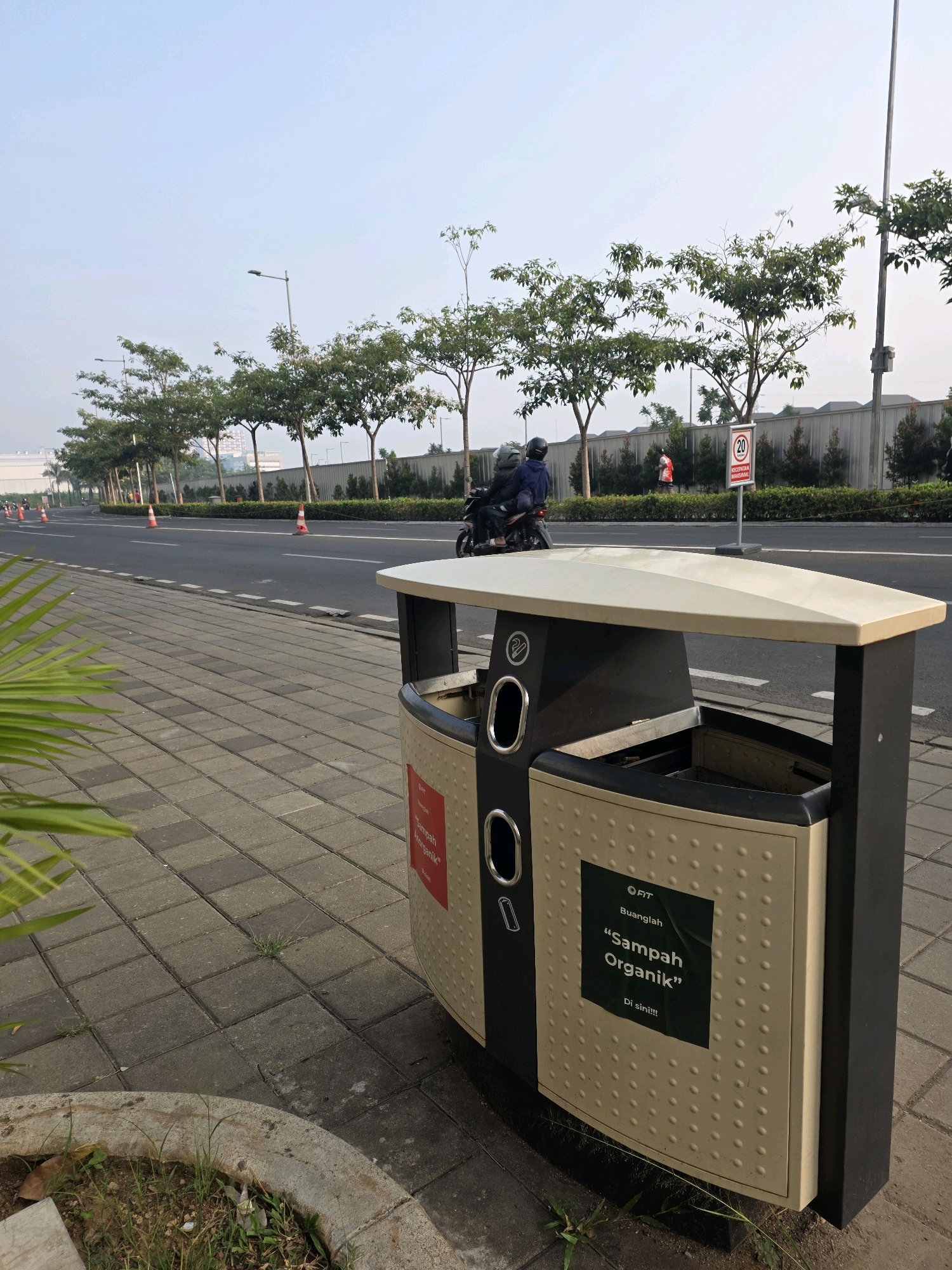 In the city of Bojongsoang, Indonesia, a street scene is captured with a focus on a segregated waste bin. The bin is beige and black with two openings. One side of the bin features a dark green sticker with white text that reads "OFIT Buanglah "Sampah Organik" Di sini!!!". To the left of the bin, a red sticker is partially visible. In the background, a road with traffic cones runs parallel to a sidewalk paved with grey square tiles. Several streetlights line the road, and a row of trees with green foliage stands behind the road. A speed limit sign indicating "20" is visible in the distance. A motorbike with two occupants, wearing helmets, is traveling on the road.
