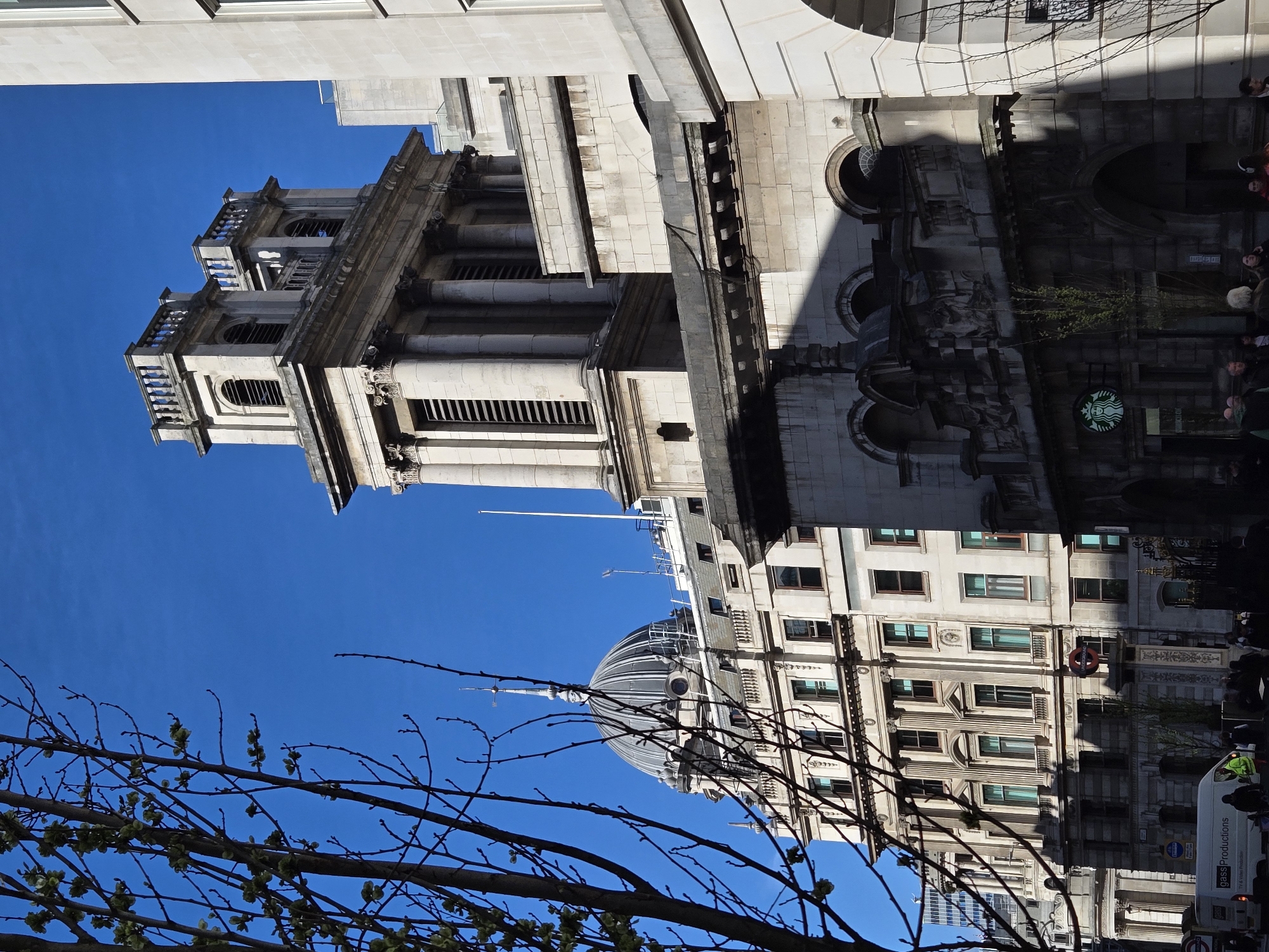 The image depicts a street scene on the Strand in London, United Kingdom, specifically looking northwest. In the foreground, partially obscuring the left side, are bare tree branches with emerging green buds. The central feature on the right is the upper portion of St Mary le Strand church, a classical stone building with a two-tiered tower featuring columns and balustrades, set against a clear blue sky. Below the church tower, on the street level, a Starbucks coffee shop logo is visible on the building's darkened facade, which features arched entrances and ornate stone carvings.
To the left and slightly behind St Mary le Strand, in the mid-ground, is Australia House, a large, multi-story stone building characterized by a prominent grey dome topped with a spired finial and weather vane. This building has numerous rectangular windows and ornate architectural details. On the street level, in front of Australia House, a red and blue London Underground roundel sign is visible on a pole. A white commercial van with "Gass Productions" text is positioned on the road, with several pedestrians visible on the sidewalk in the lower portion of the image. The scene is illuminated by bright sunlight, casting strong shadows on the buildings.