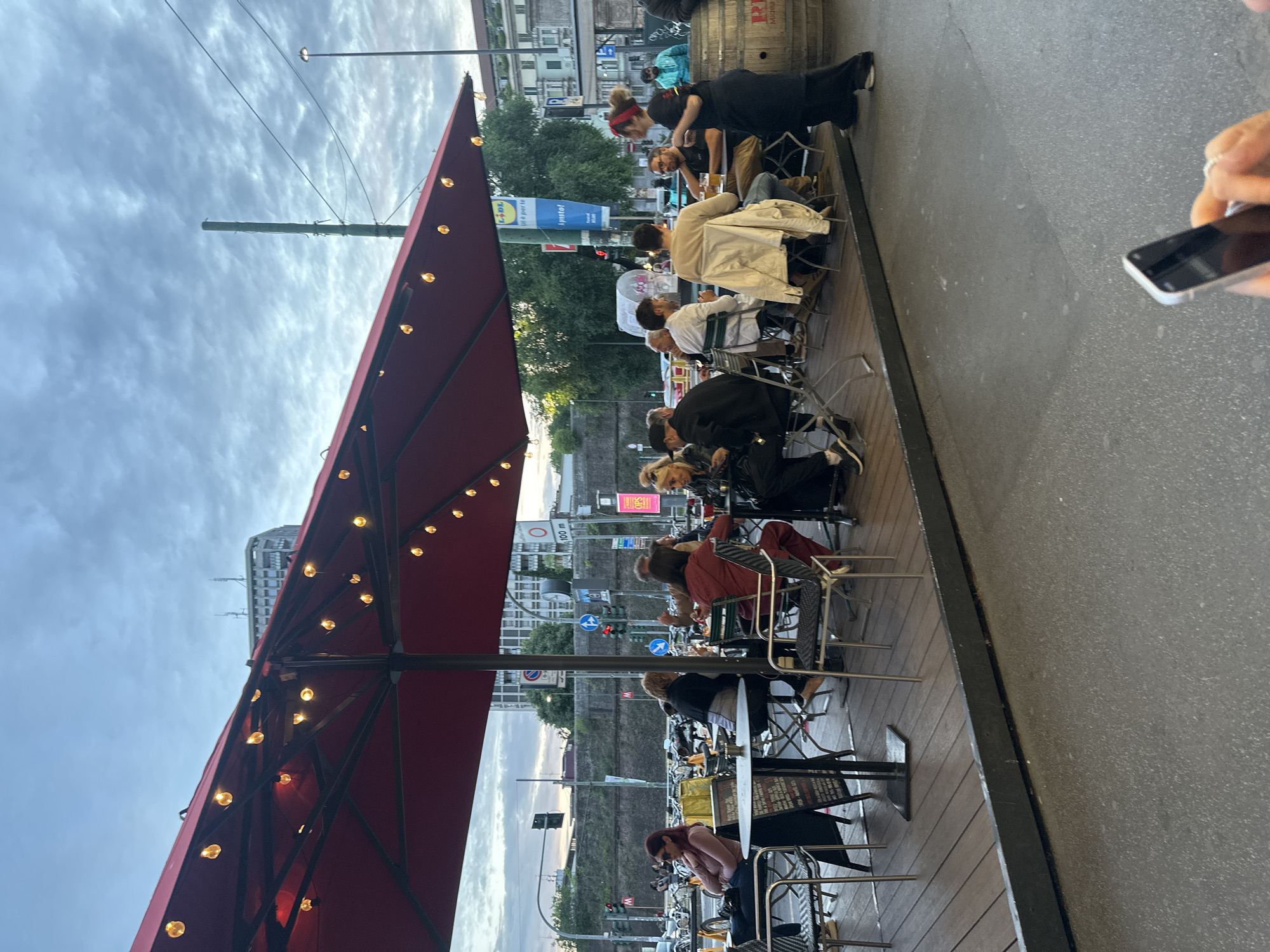 The image depicts an outdoor restaurant setting in Milan, Italy. A large, red umbrella with string lights provides shade over multiple tables occupied by patrons. People are seated at tables, interacting with each other and engaging in dining activities. A server is attending to a table near a wooden barrel. Street signs and advertisements are visible in the background. Cars and pedestrians are present on the street beyond the seating area. The sky is overcast. A hand holding a mobile phone is visible in the right foreground. The location is near a Lidl grocery store.