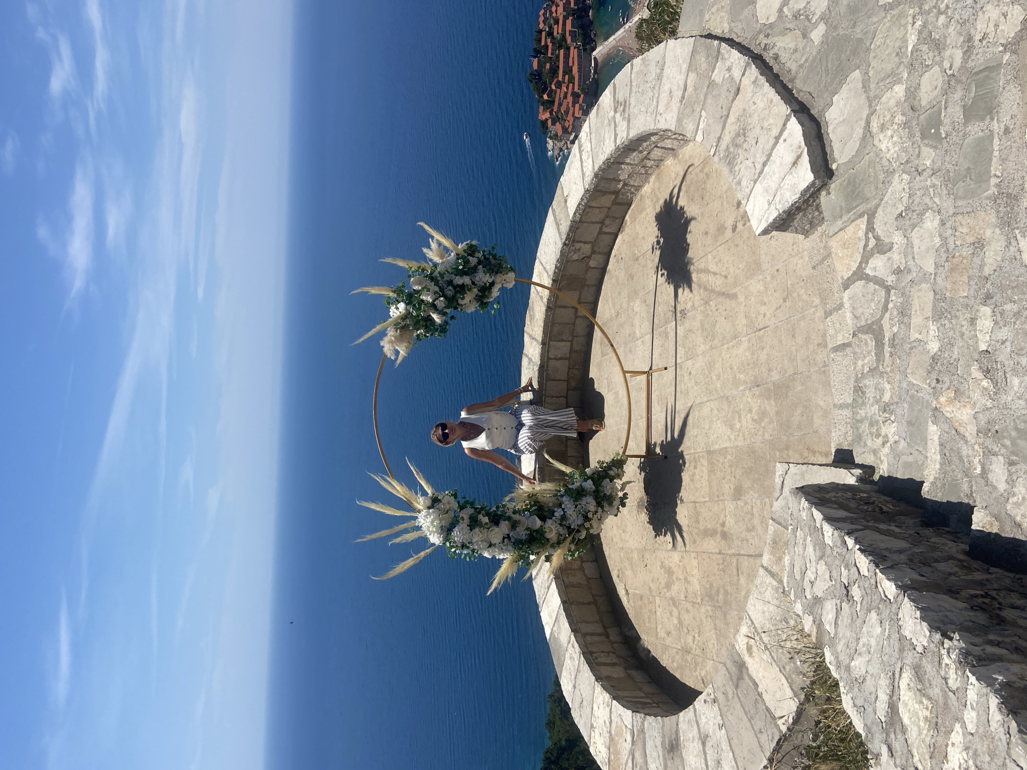 A woman wearing sunglasses, a white sleeveless top, and striped pants sits on a stone wall overlooking the Adriatic Sea. She is positioned within a circular golden arch decorated with white flowers and feathery reeds. The arch is set on a paved circular platform with stone steps leading down and to the left. In the background, the blue sea meets a hazy sky, and a small island with red-roofed buildings, likely Sveti Stefan, is visible to the right. The scene is bathed in bright sunlight, casting shadows on the platform.