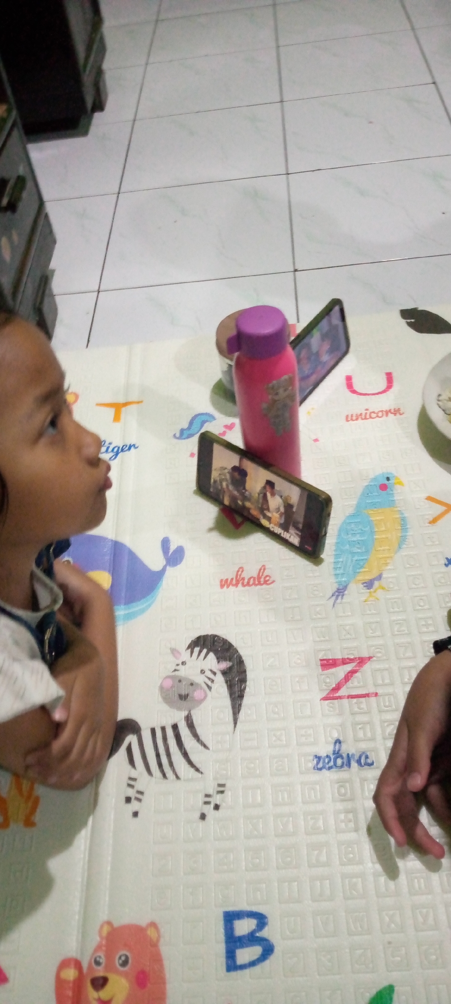 The image captures an indoor scene on a white mat with colorful educational graphics of animals and letters. In the foreground, a young child with dark hair and brown eyes is looking upwards, with their mouth slightly open as if speaking or making a sound. The child is wearing a white shirt with a blue collar. To the right of the child, a pink water bottle with a purple lid stands upright. Two smartphones are placed on the mat, one in front of the water bottle and another behind it, both displaying videos. The mat is patterned with a variety of elements, including a blue whale, a zebra, and the word "unicorn." In the background, a tiled floor is visible, and a dark-colored furniture item, possibly a dresser or cabinet, is present. The lighting suggests an indoor setting with artificial or ambient light. The image was taken in Sumberwono, Indonesia.