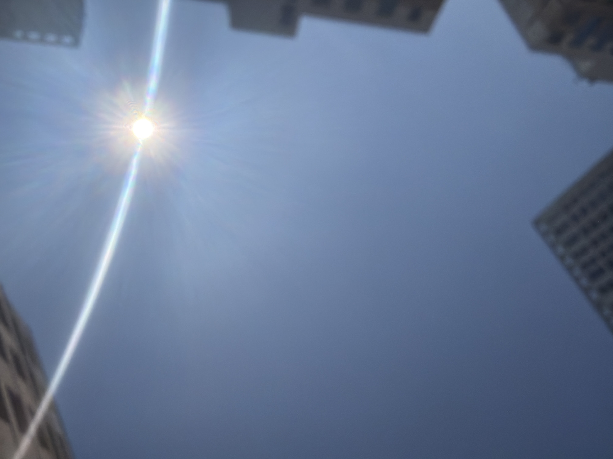 The image depicts a low-angle view of a clear blue sky with the sun prominently visible, emitting strong lens flare and a rainbow-like halo effect. Several out-of-focus buildings, characteristic of an urban environment, are visible at the edges of the frame, suggesting a city setting. The architectural style of the buildings, with their geometric patterns and visible window structures, is consistent with modern high-rise structures found in cities like Yekaterinburg, Russia. The overall impression is that of a bright, sunny day in an urban landscape, viewed from ground level looking upwards. There are no people or identifiable actions captured in the image.