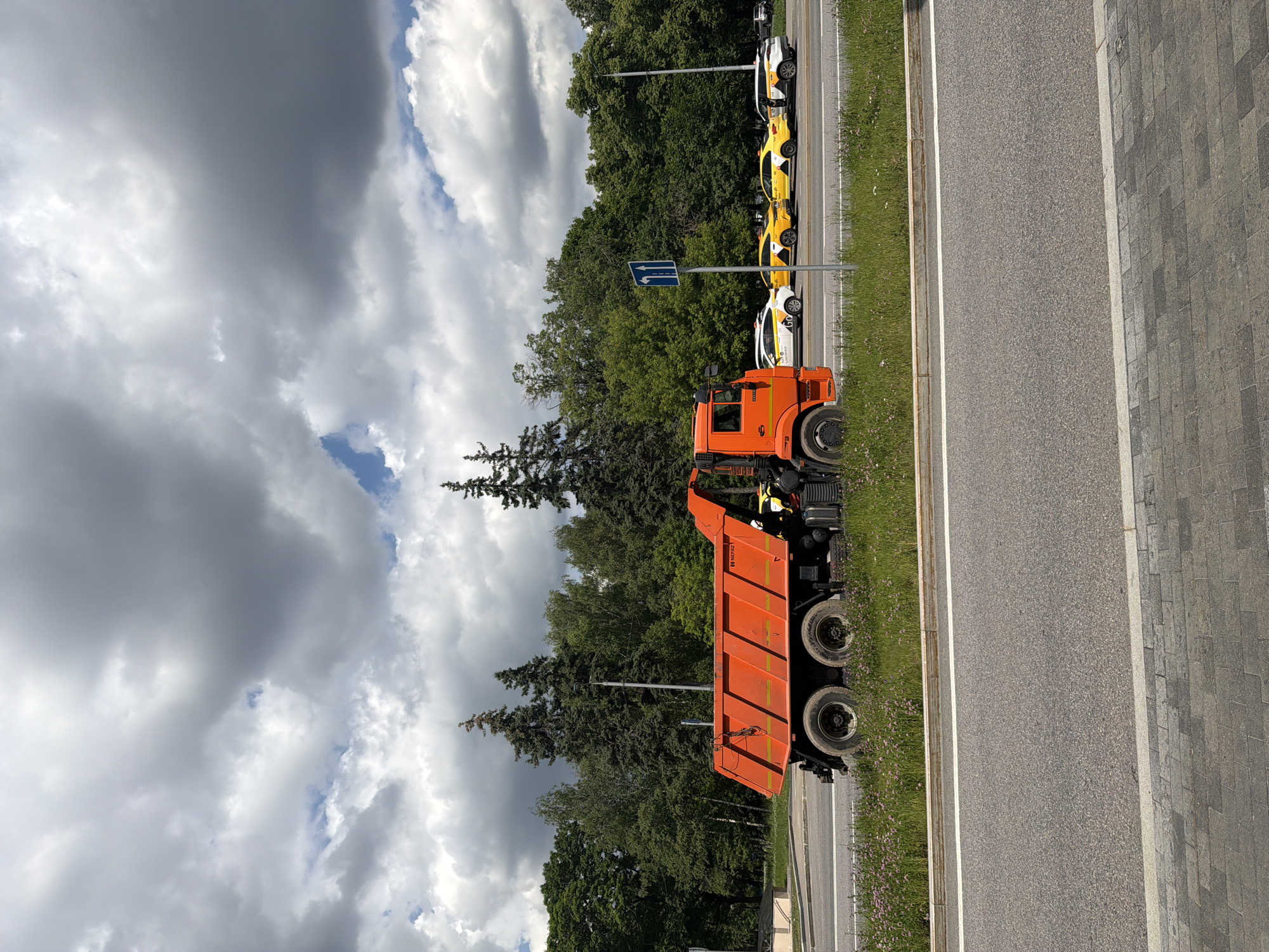 An orange dump truck is parked on the side of a road, with a line of yellow taxis behind it. The dump truck is facing left and is partially obscured by a patch of tall grass and flowers. The yellow taxis are all facing right and appear to be waiting in traffic. Behind the taxis, a dense forest of green trees stretches across the background. The sky is filled with dramatic, heavy clouds, suggesting an overcast day. The road is paved with asphalt, and a sidewalk made of grey and tan pavers is visible in the foreground. The location is a city road lined with trees and a grassy median.