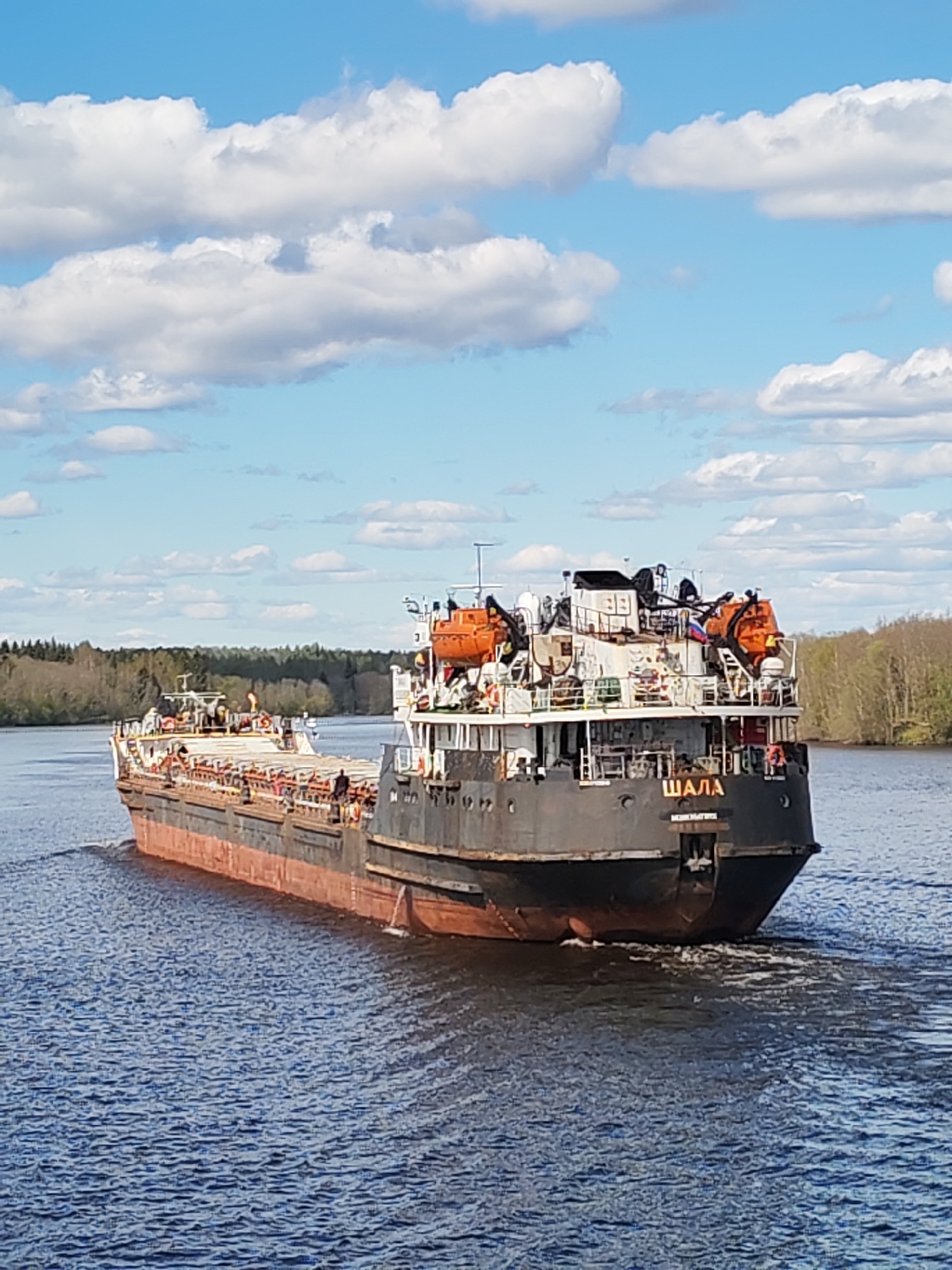 The image shows a large, older cargo ship on a waterway. The ship, named "ШАЛА" (Shala) in Cyrillic script, is dark gray and red. The stern displays "МОСКОВСКИЙ" (Moscowsky) and "ИВАНОВ БОР" (Ivanov Bor), indicating a connection to the city of Ivanov Bor in Russia. The ship is moving away from the viewer, creating a wake in the water. The deck of the ship carries cargo and lifeboats. A Russian flag is visible. The surrounding environment includes a blue sky with scattered clouds, and tree-lined banks on both sides of the waterway.