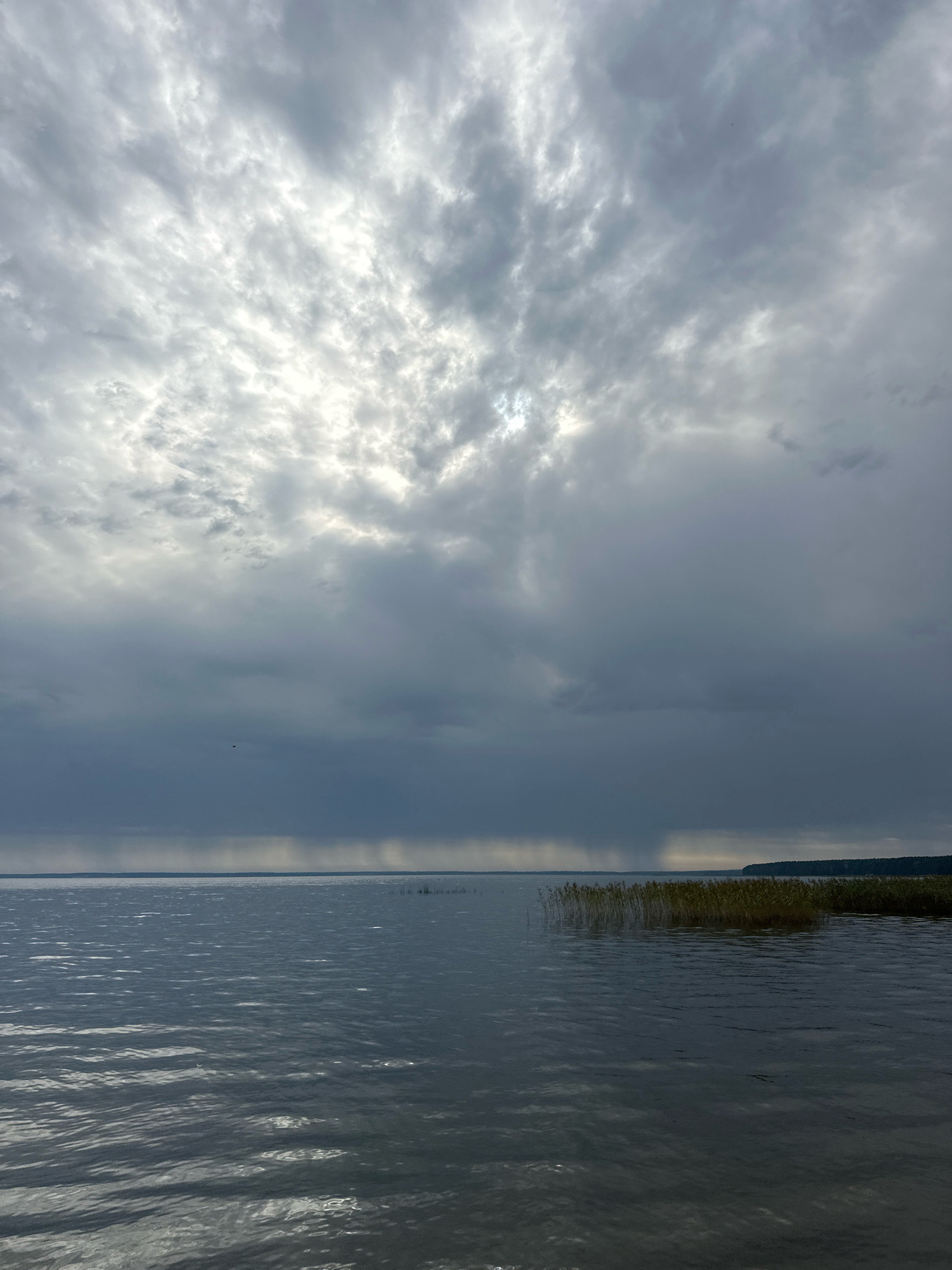 This is an outdoor, full shot of a cloudy sky over a large body of water. The sky is filled with gray, heavy clouds, with streaks of sunlight breaking through in the upper left quadrant. The water is dark and choppy, reflecting the cloudy sky. In the distance, a thin line of trees or vegetation can be seen along the horizon. The scene appears to be a natural landscape, possibly a lake or a wide river, with no human presence or structures visible. The location is identified as Нарацкі сельскі Савет, Belarus.