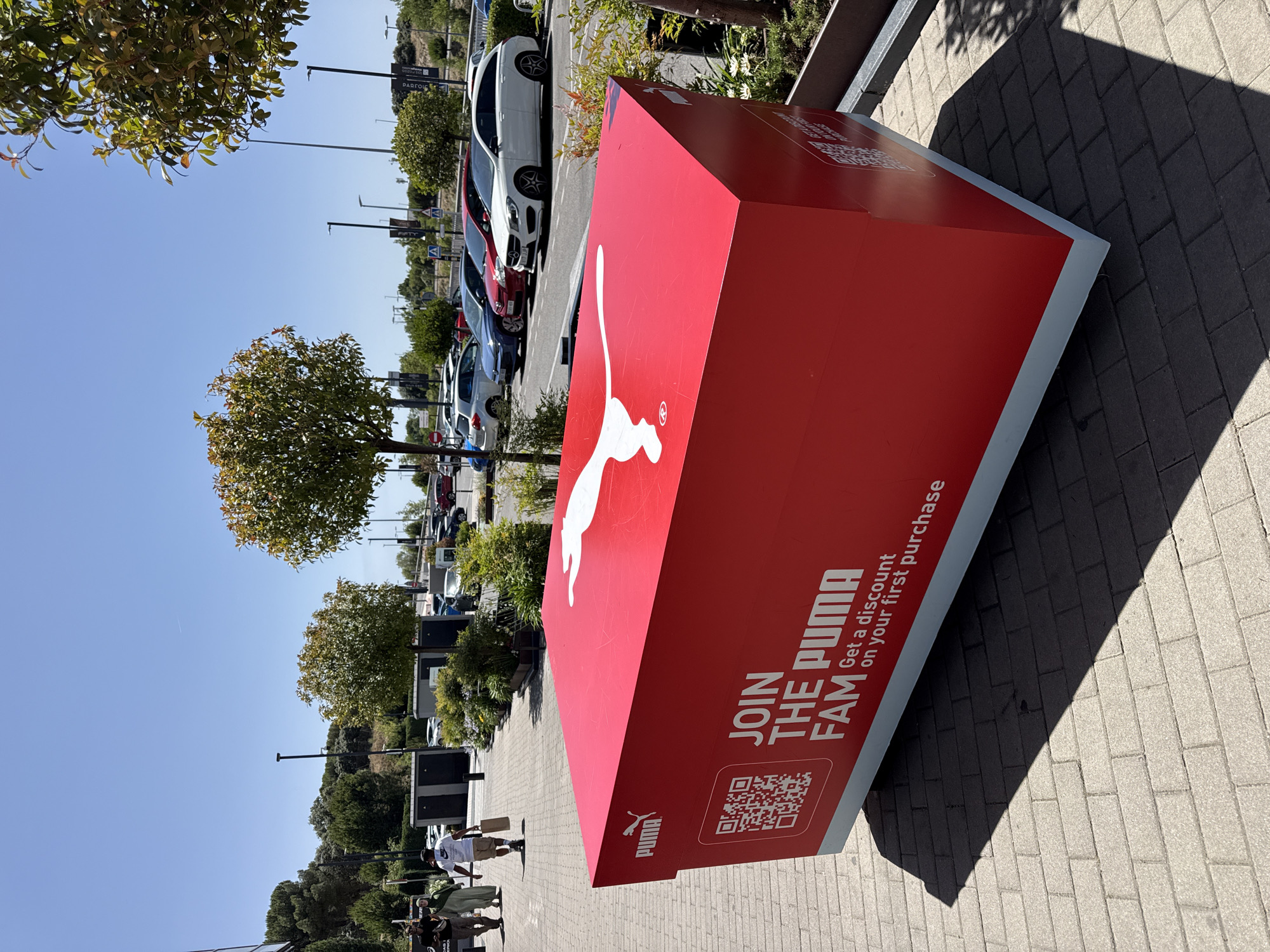 A bright, sunny day is depicted in this outdoor scene in Las Matas-Pinar-Monte Rozas, Spain. The foreground is dominated by a large, red, rectangular object resembling an oversized Puma shoebox. The box has the Puma logo on its side and top, and text that reads "JOIN THE PUMA FAM Get a discount on your first purchase." A QR code is also visible on the side of the box. The object rests on a grey paved surface with a distinct shadow cast to the left. In the background, a parking lot filled with cars stretches into the distance. Several trees with green foliage are scattered throughout the scene. A few individuals are walking in the background on a paved walkway. The sky is clear and blue.